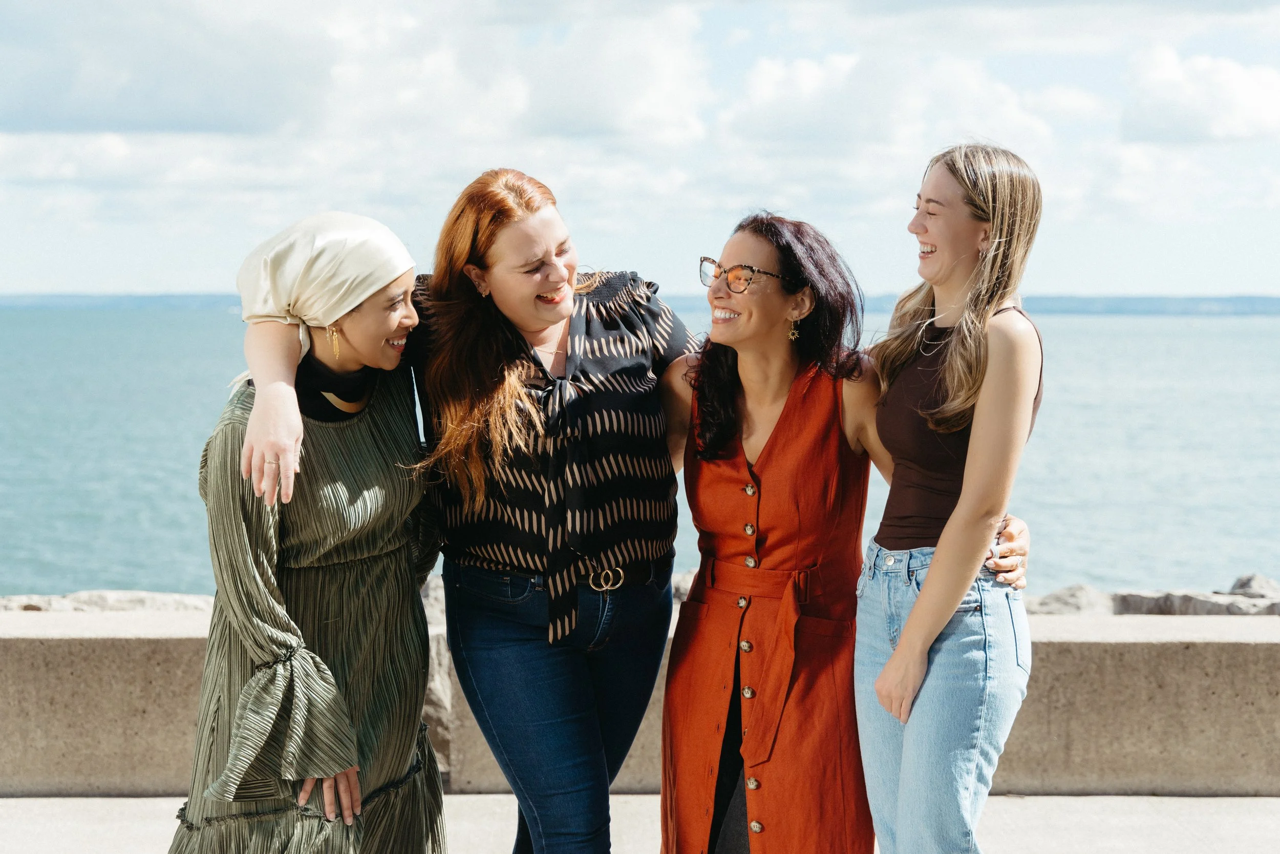 Five women smiling and hugging by the water on a sunny day.