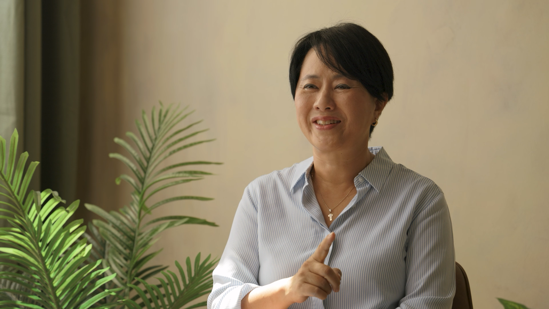 A woman smiling and pointing her finger in front of a beige wall and green plants.