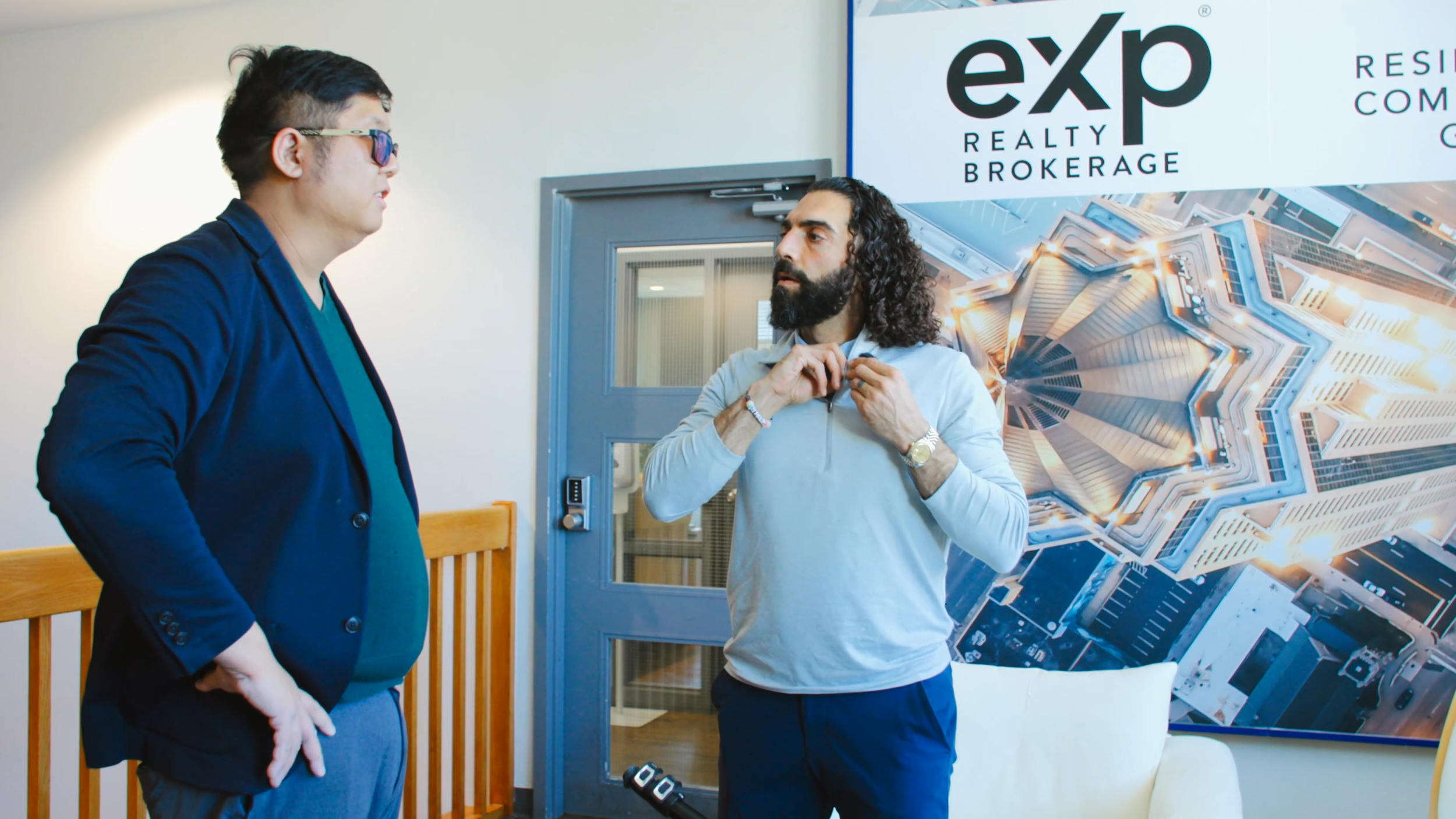 Two men having a conversation indoors, with a large Expo Realty Brokerage sign and a cityscape mural behind them.