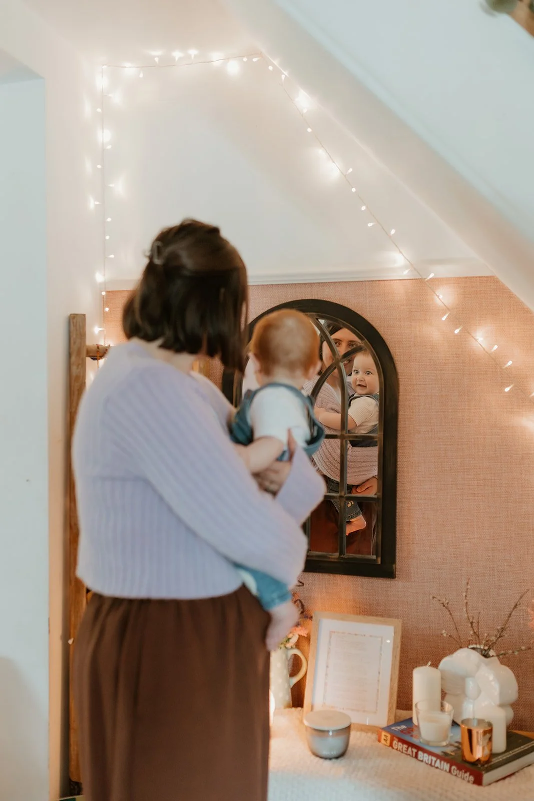A woman holding a baby looking into a mirror with fairy lights hanging from the ceiling, cozy decor and books on a table.