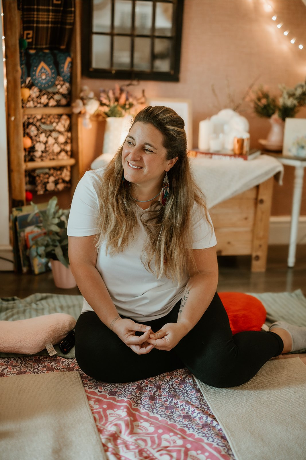 Woman with long, wavy hair, smiling, sitting cross-legged on a patterned mat in a cozy room with wooden furniture, plants, and decorative items.