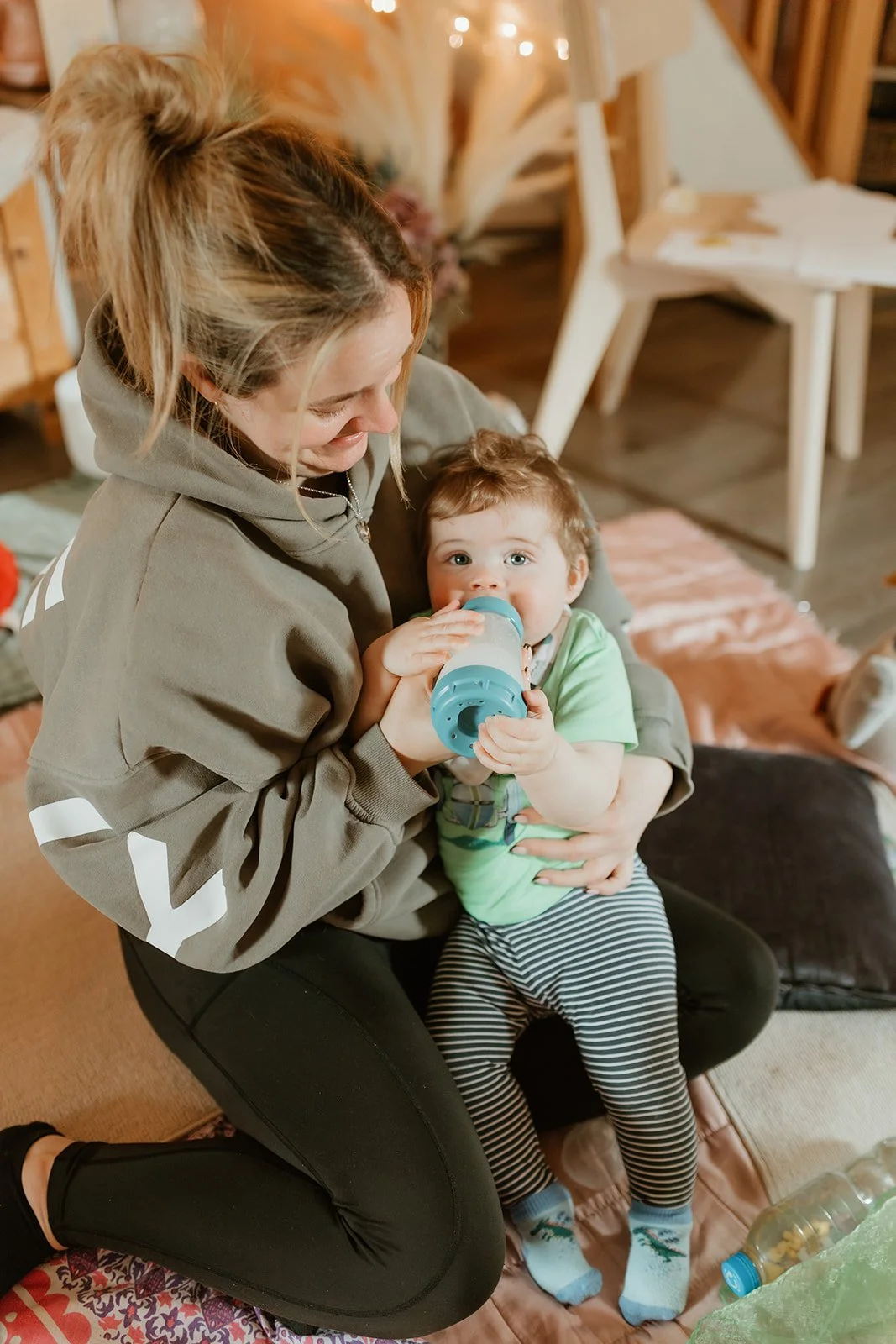 A woman is sitting on the floor holding a young child while feeding him from a bottle. The woman has blonde hair tied up in a messy bun and is wearing a gray hoodie. The child is wearing a green shirt, black and white striped pants, and socks, and is looking directly at the camera.