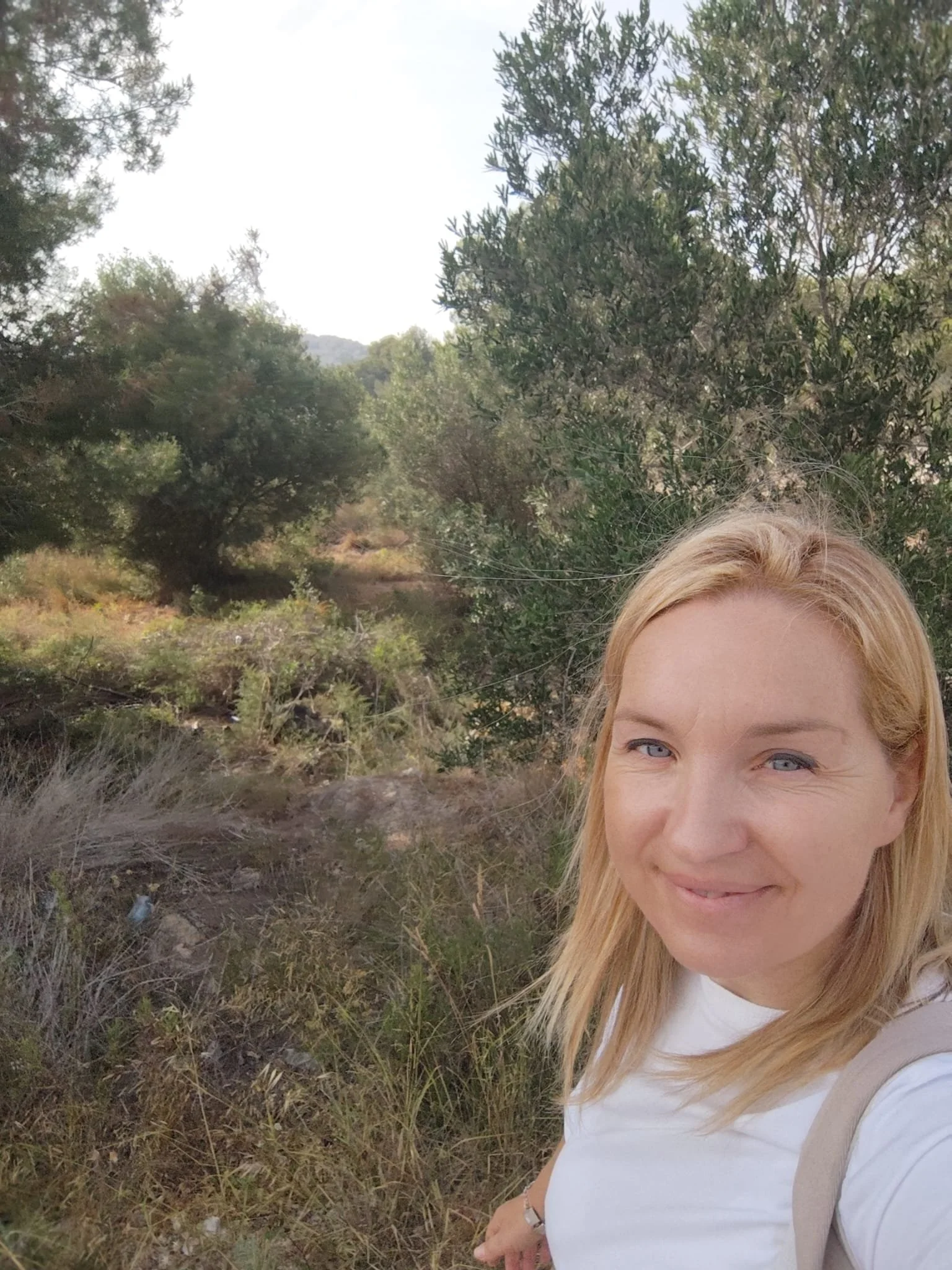 A woman with blonde hair and blue eyes taking a selfie outdoors in a pasture with trees and bushes in the background.