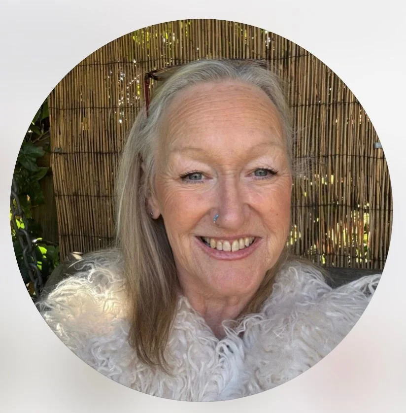 Smiling woman with long blonde hair and a nose piercing, wearing a white fluffy top, sitting outdoors with a bamboo fence in the background.
