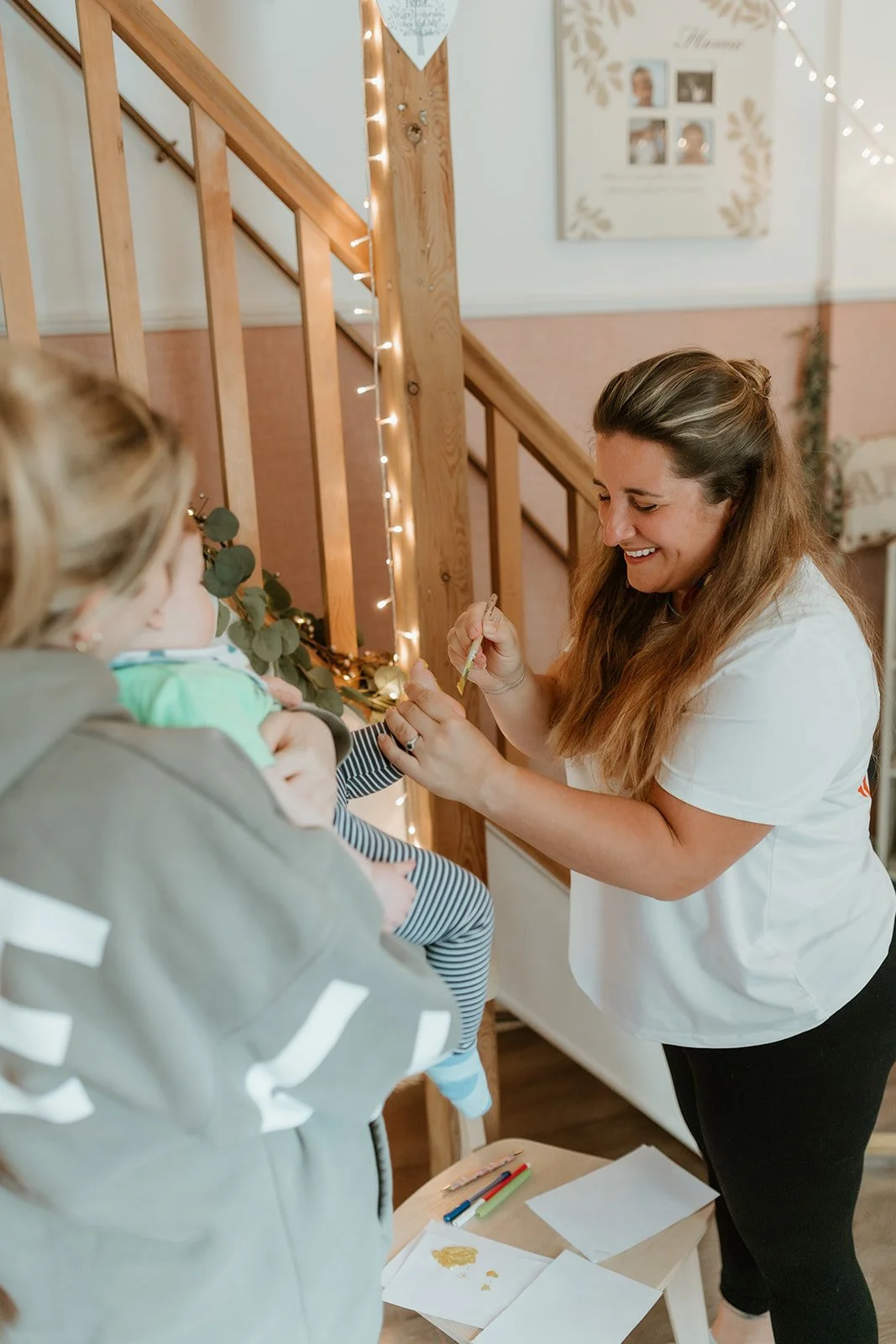 A woman is giving a vaccination shot to a child, who is being held by an adult. The woman is smiling, and they are in a cozy indoor space decorated with string lights and artwork on the wall.
