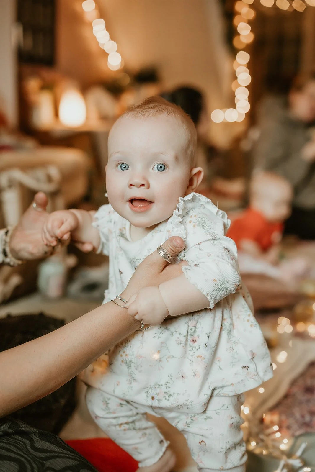 A smiling baby girl with blue eyes, wearing a white floral dress, is being held by an adult at a festive gathering with warm lights in the background.