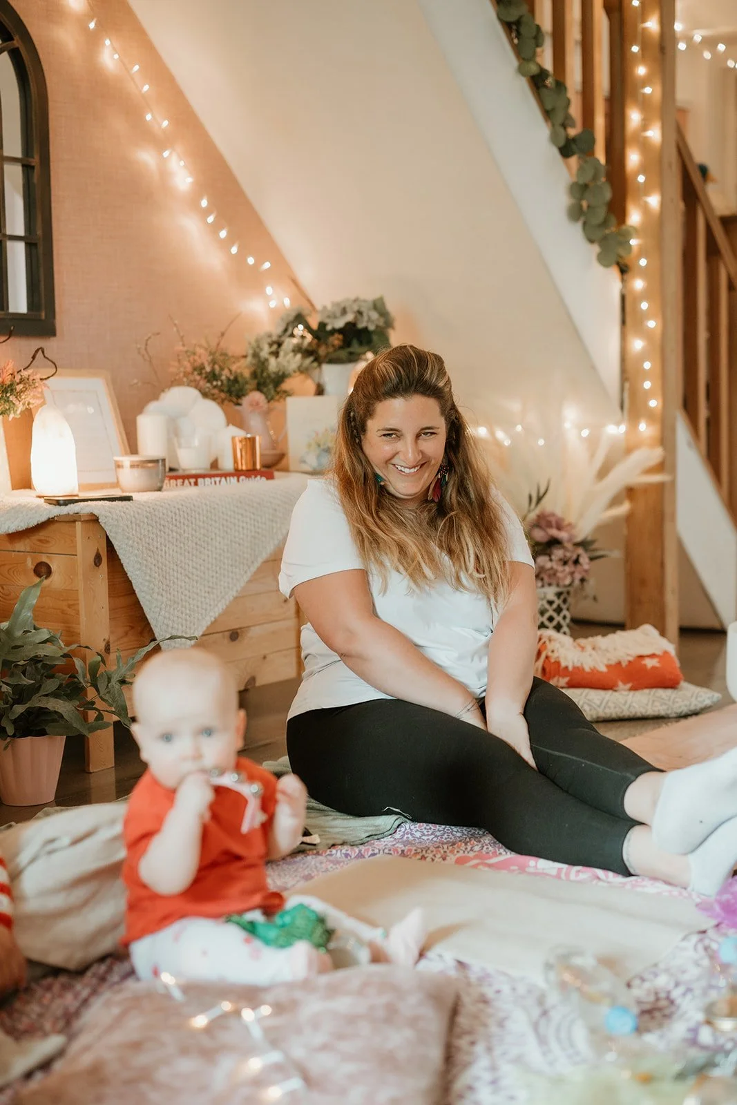 A woman sitting on the floor smiling, with a baby in front of her, in a cozy decorated living space with string lights, plants, and decorative objects.