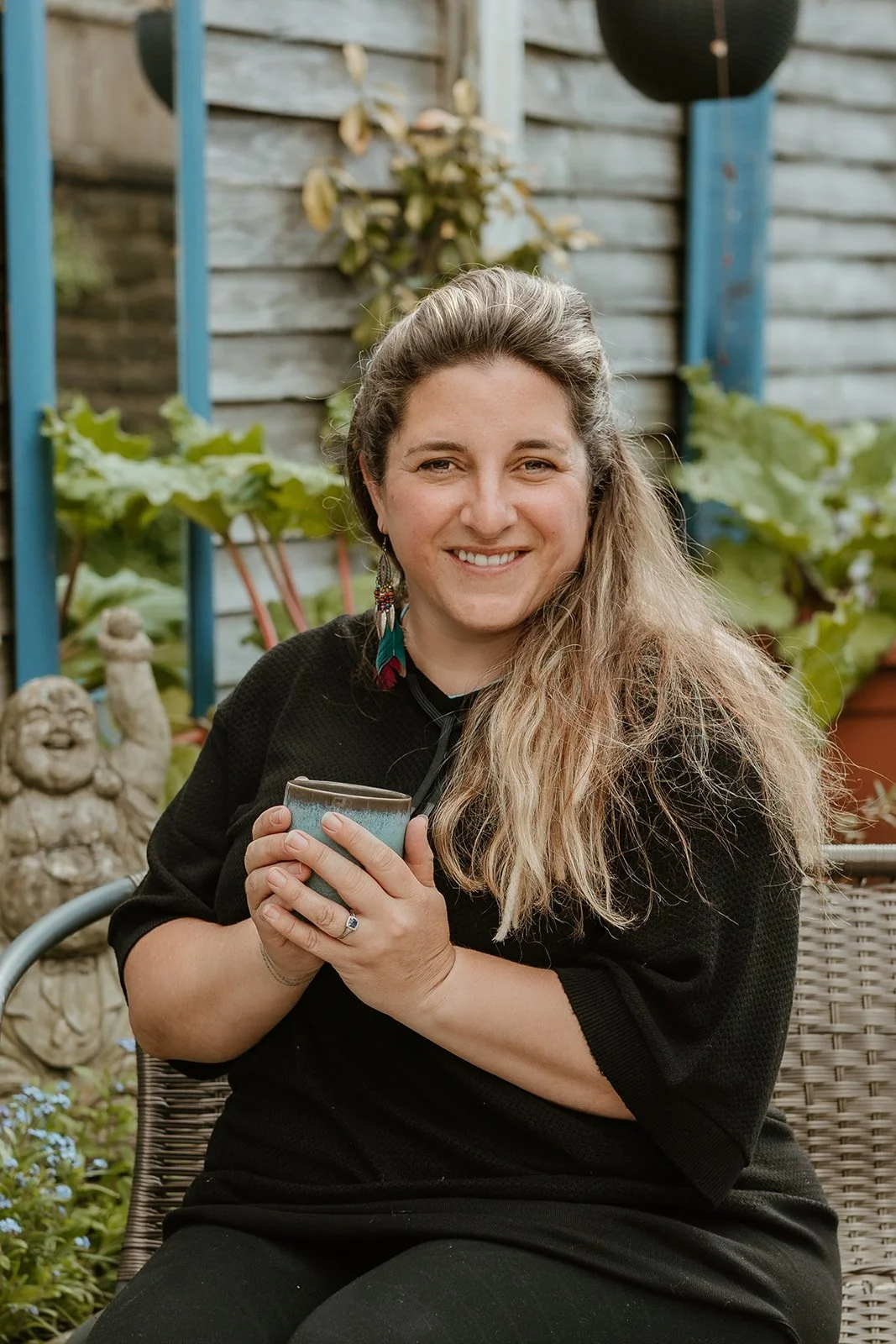 A woman with long, light brown hair smiling and holding a mug, sitting outdoors in a garden. She is wearing black. There are plants, a Chinese laughing Buddha statue, and gardening tools in the background.