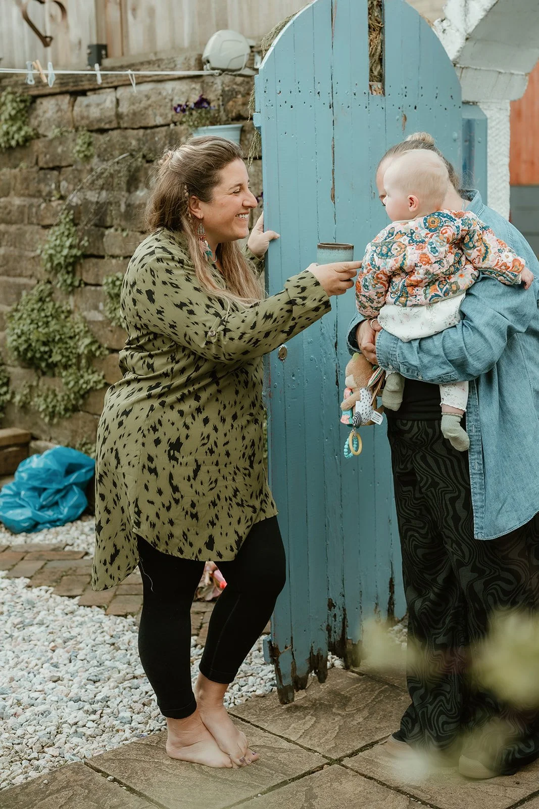 A woman smiling and holding a mug while standing barefoot outside near a blue wooden gate, interacting with a person holding a baby dressed in a colorful floral jacket.