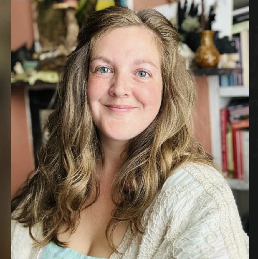 A woman with long, wavy light brown hair and blue eyes, smiling at the camera. She is wearing a cream-colored textured top. The background features a bookshelf with books and decorative items.