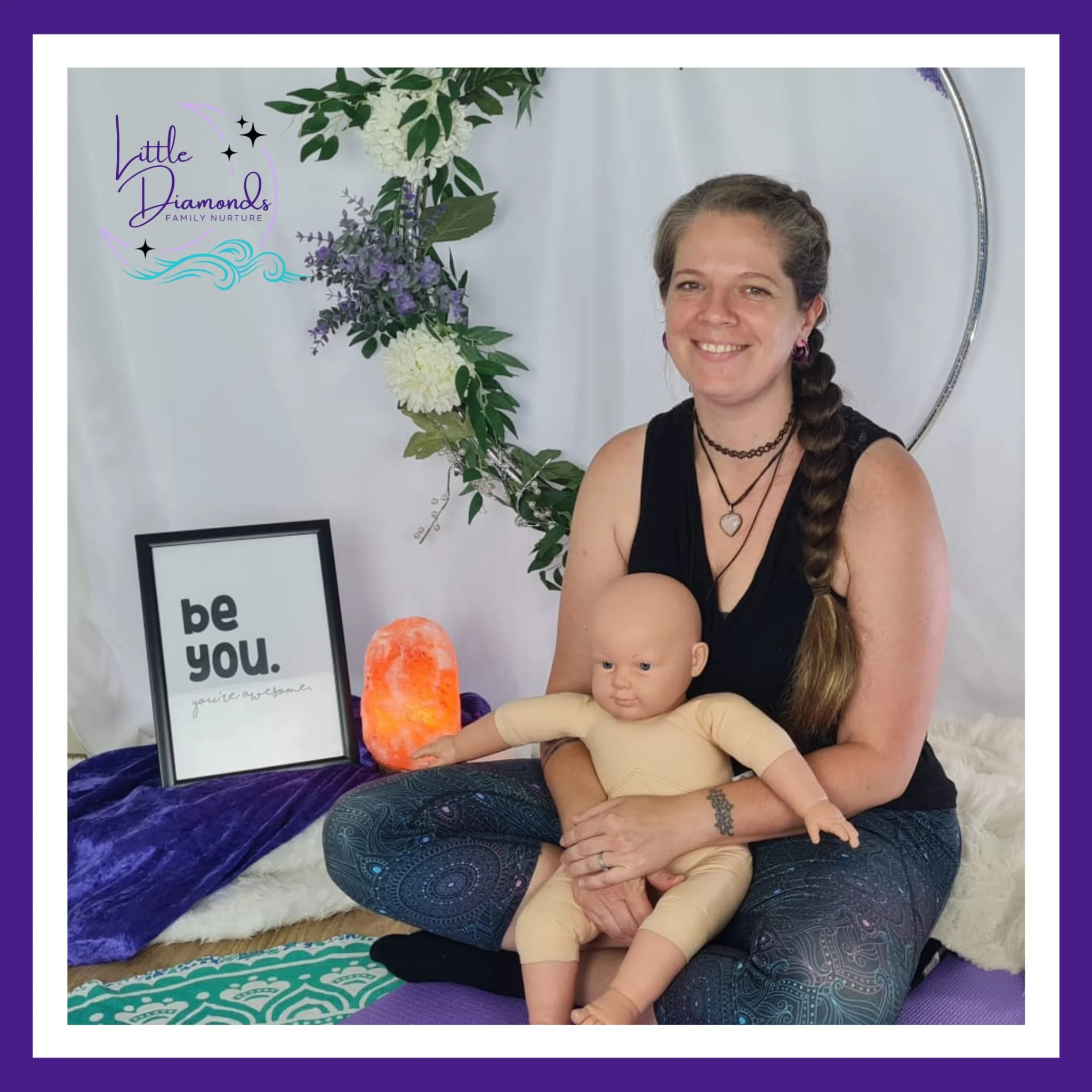 A woman with long braided hair and tattoos on her hand is sitting on the floor with a young child, both smiling. Behind them are decorative floral arrangements and framed posters, one of which says 'be you'.