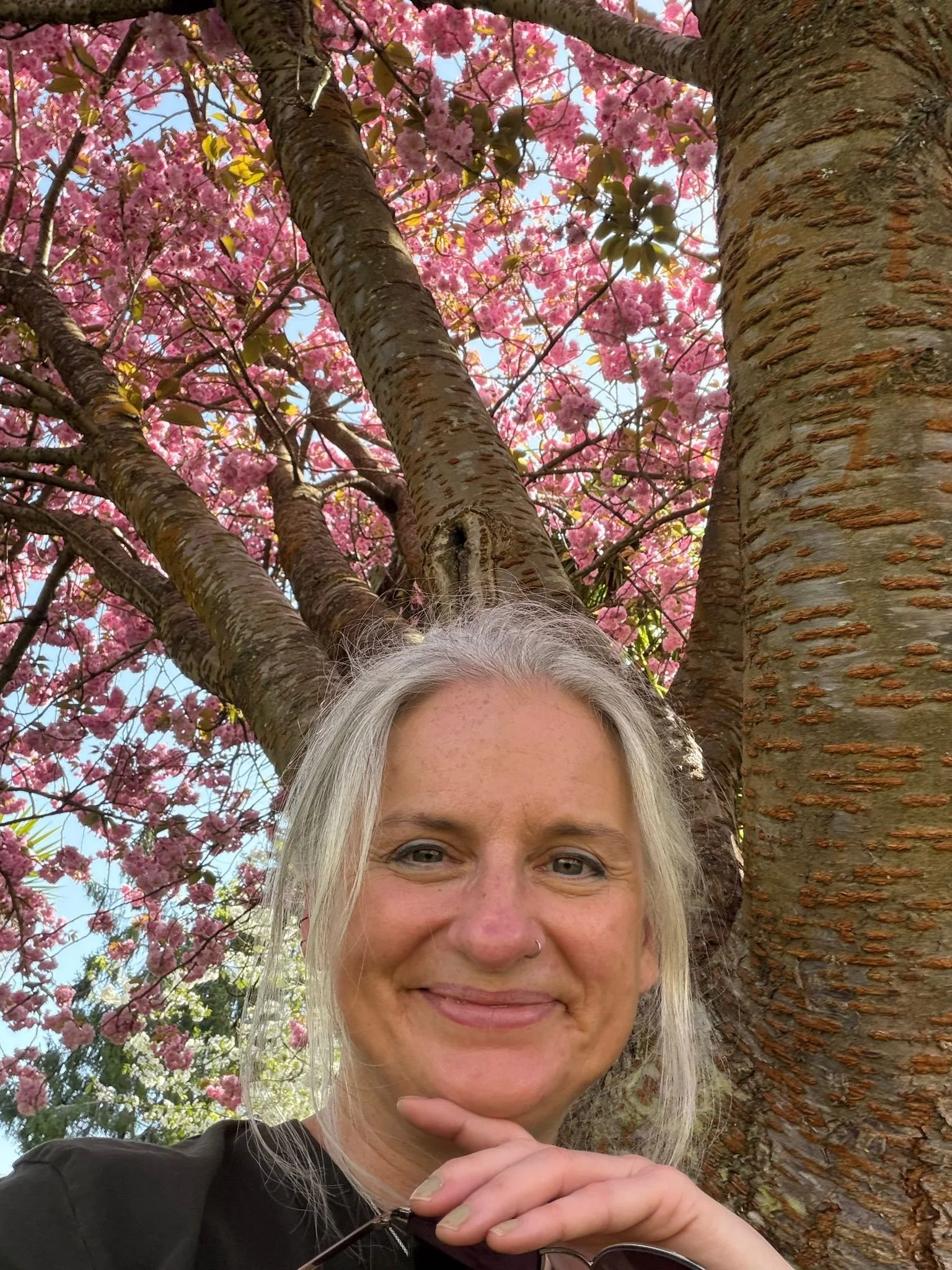 A woman with light gray hair, smiling, standing in front of a flowering pink tree. The sky is visible through the branches and blossoms.