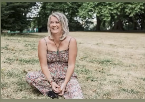 A smiling woman with gray hair sitting on the grass in a park, wearing a floral dress.
