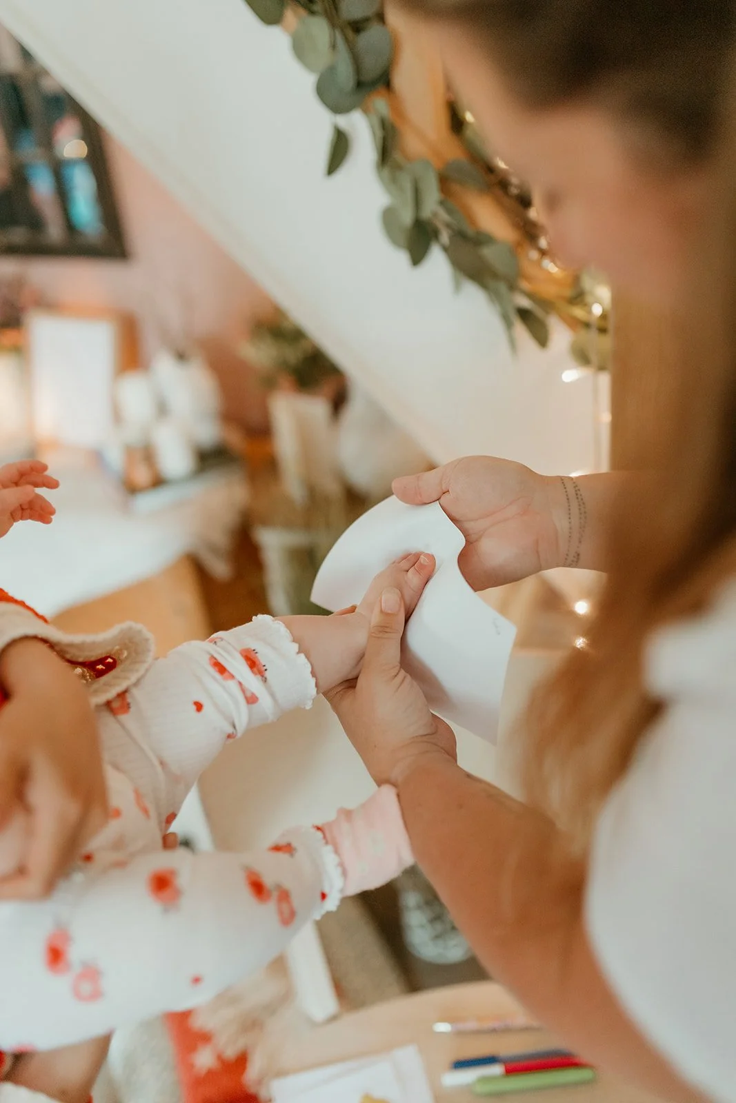 A woman gives a tissue to a child with a holiday pajama outfit, in a cozy decorated living room with candles and greenery