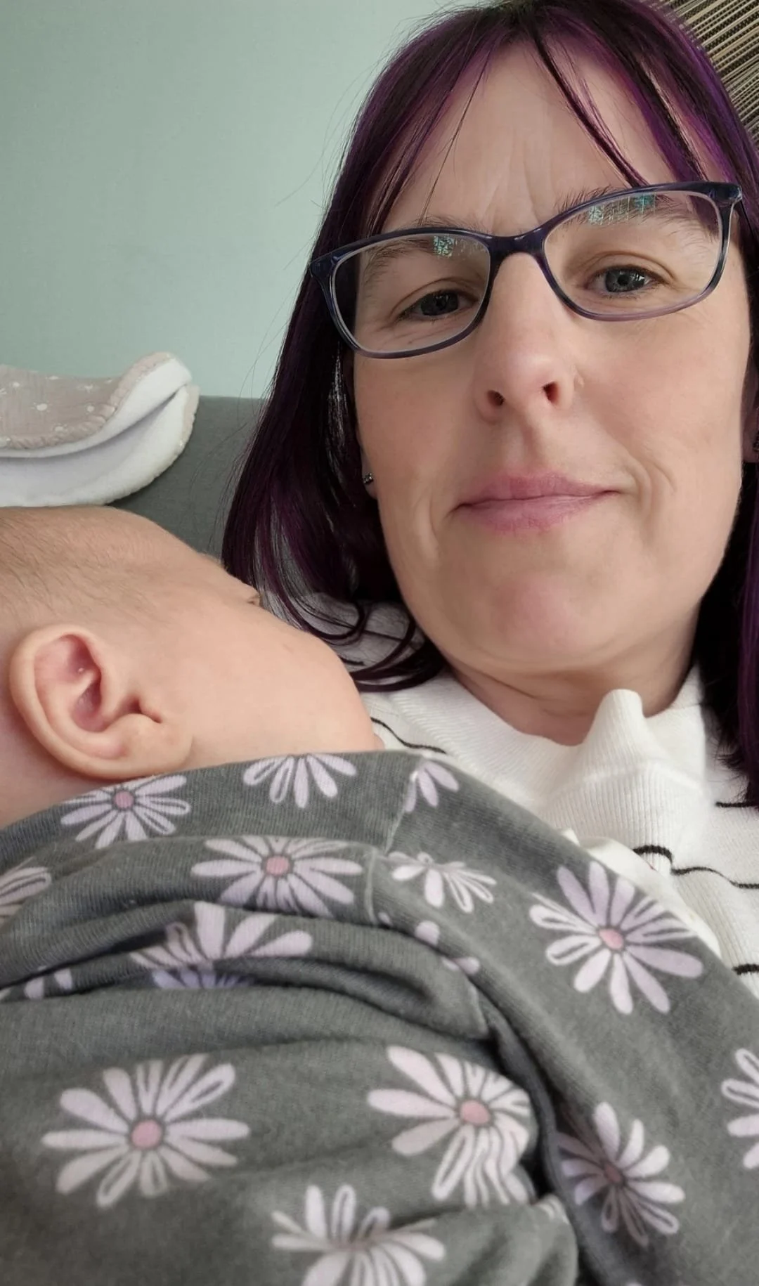 A woman with purple hair and glasses is holding a sleeping baby with a floral-patterned shirt, close to her face, in a cozy setting.