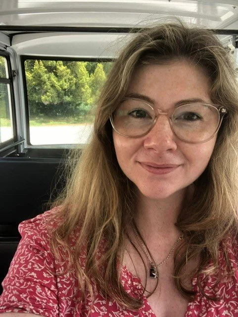 A woman with light brown hair, glasses, and a red patterned shirt smiling while sitting on a vehicle with a window showing green trees outside.