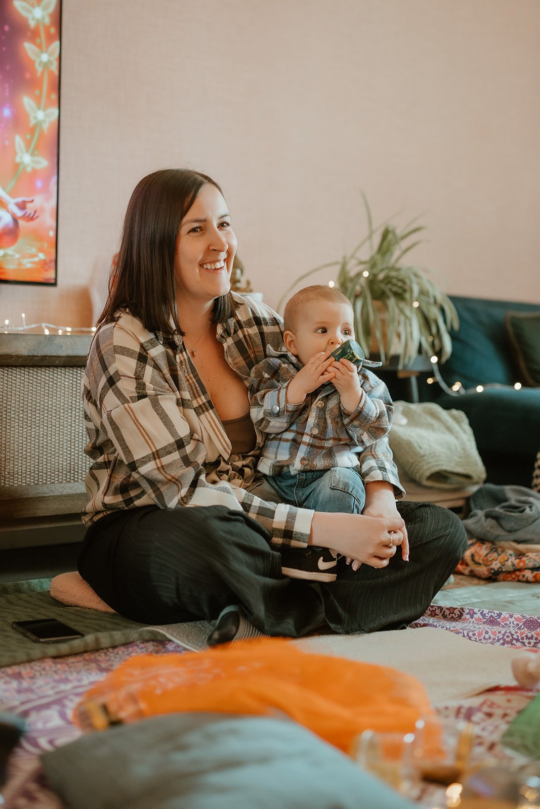 A smiling woman and a child sitting on a floor decorated for Christmas, with holiday lights and a TV in the background. The woman is holding the child, who is drinking from a cup.
