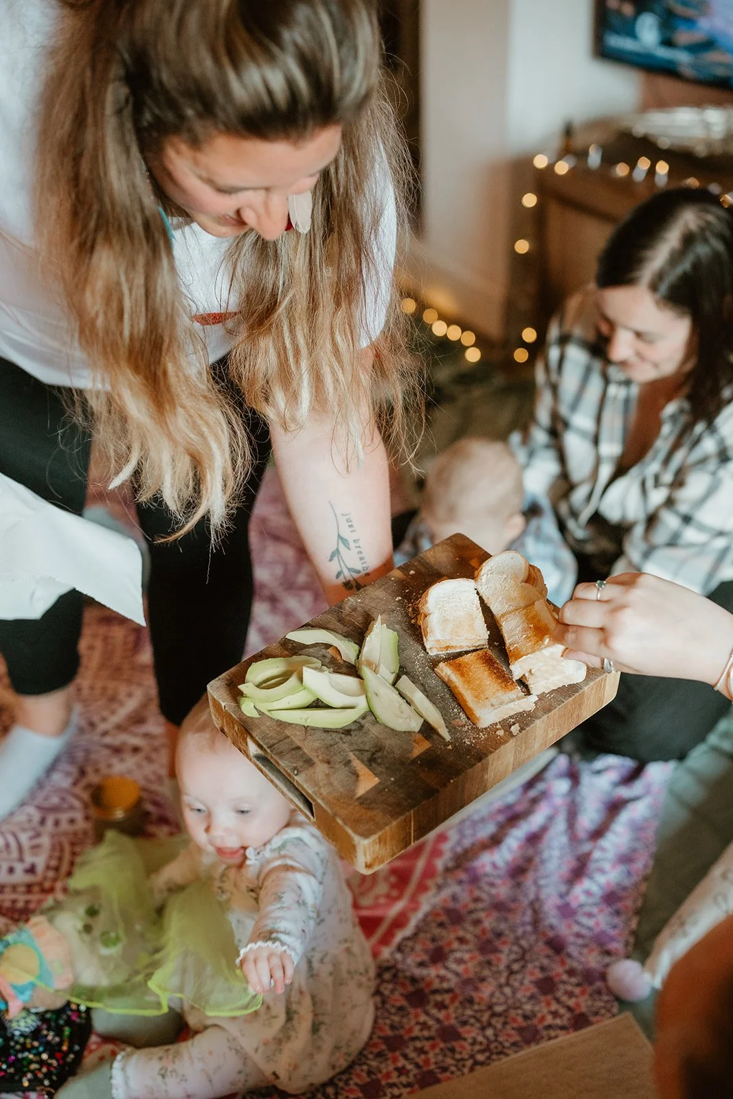A woman with long hair holding a cutting board with slices of bread and onion, two women and a child in the background, and a baby sitting on the floor in front, with Christmas lights in the background.