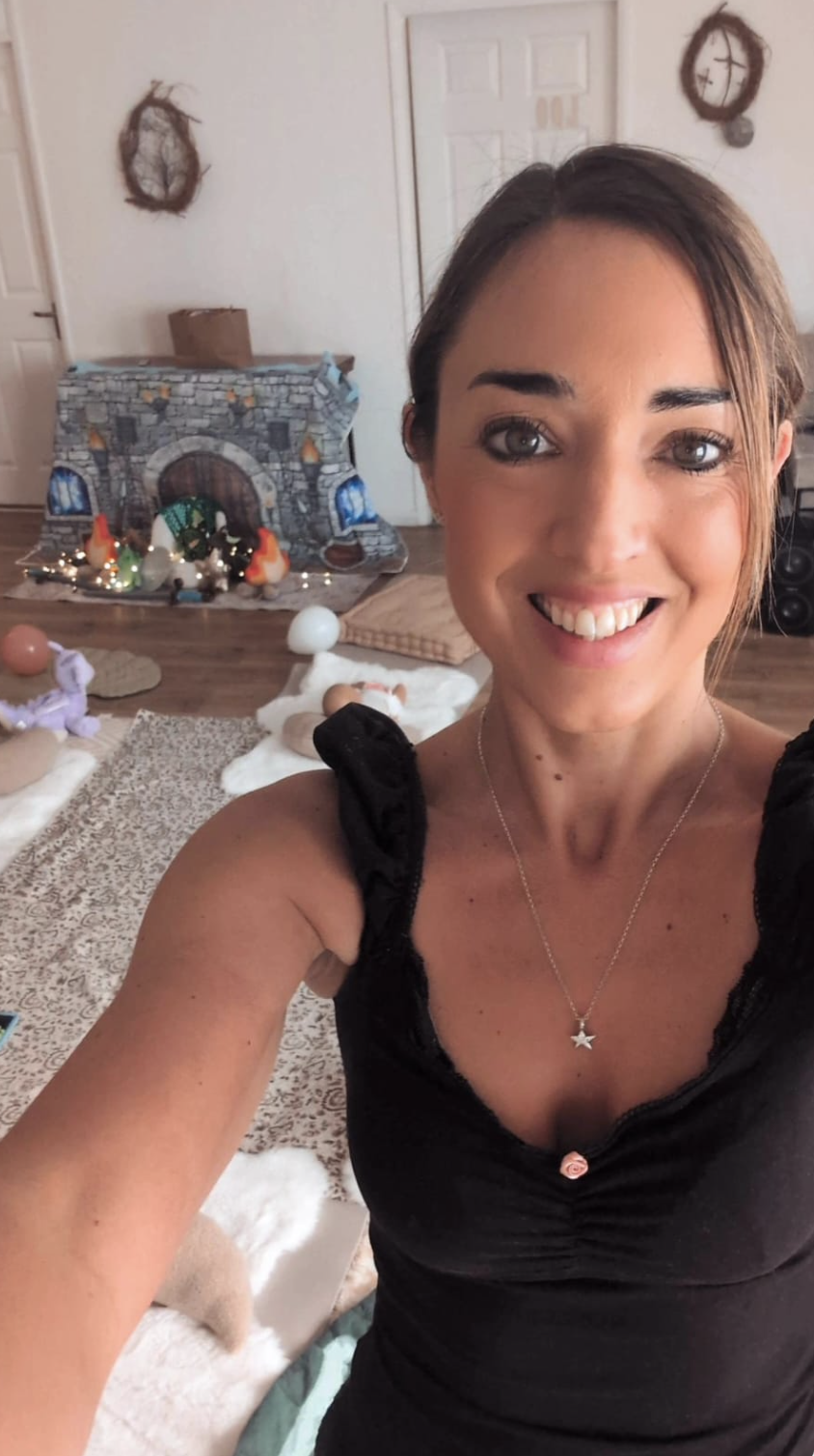 A woman taking a selfie in a room decorated for Christmas. In the background, there is a fireplace setup with festive decorations, including toy people, lights, and a snowy scene. The woman is smiling, wearing a black top with puffed sleeves and a star necklace.