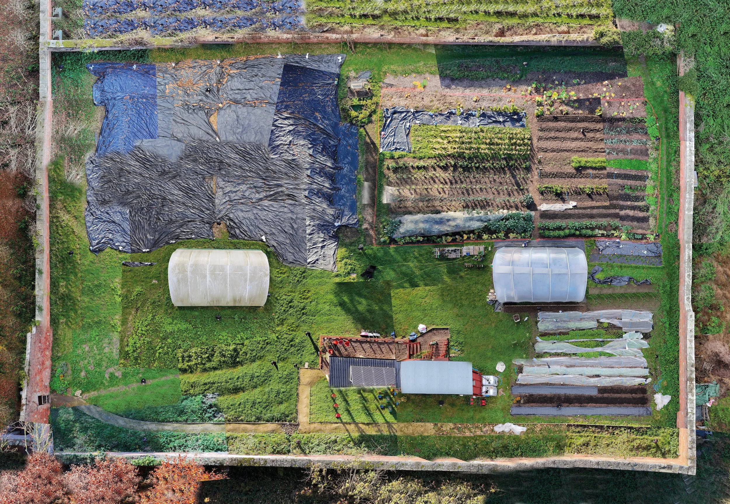 An aerial view of a backyard garden with divided sections, including greenhouses, planting beds, and a cover over part of the garden, surrounded by a fence and trees.