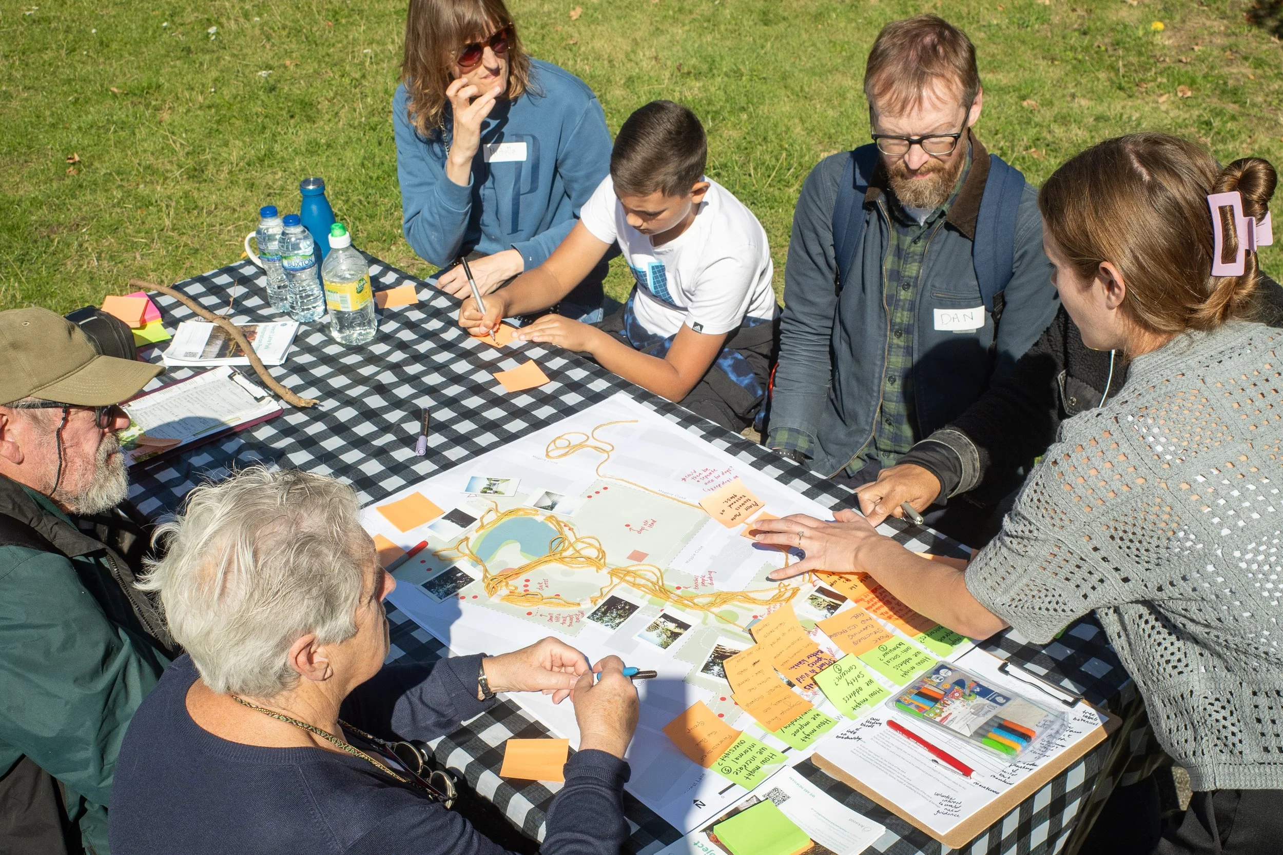 Group of people sitting and standing around a table outdoors, working on a project with maps, notes, and markers, on sunny day.
