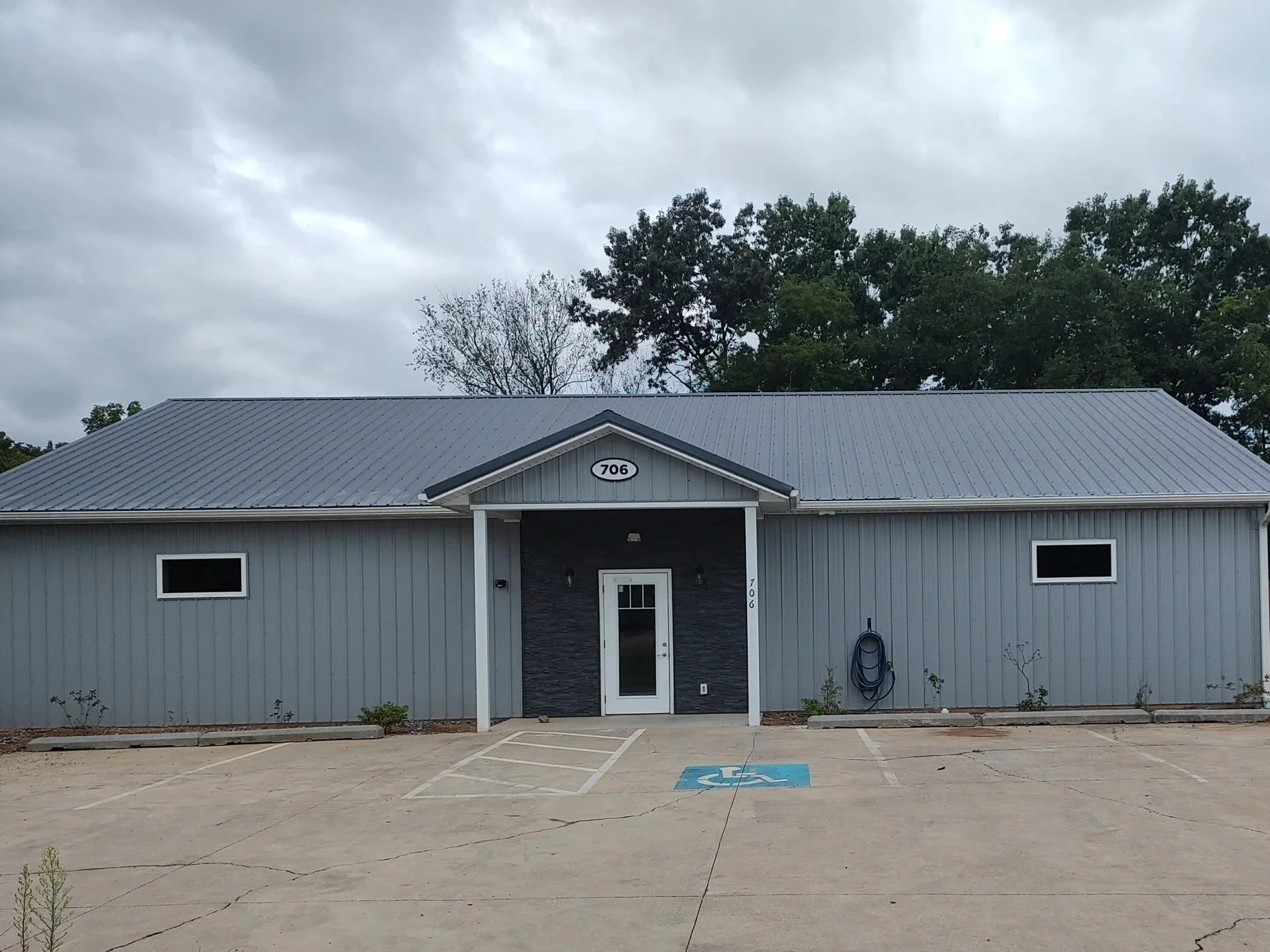 A grey commercial building with a metal roof, parking lot with a designated handicapped parking space marked with a blue symbol, and a blue hose mounted on the wall, under a cloudy sky.