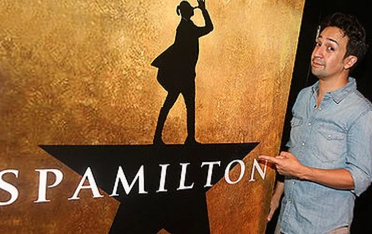 A man (Lin-Manuel Miranda) standing next to a sign featuring a silhouette of a woman standing on a star with the name 'SPAMILTON' on it.