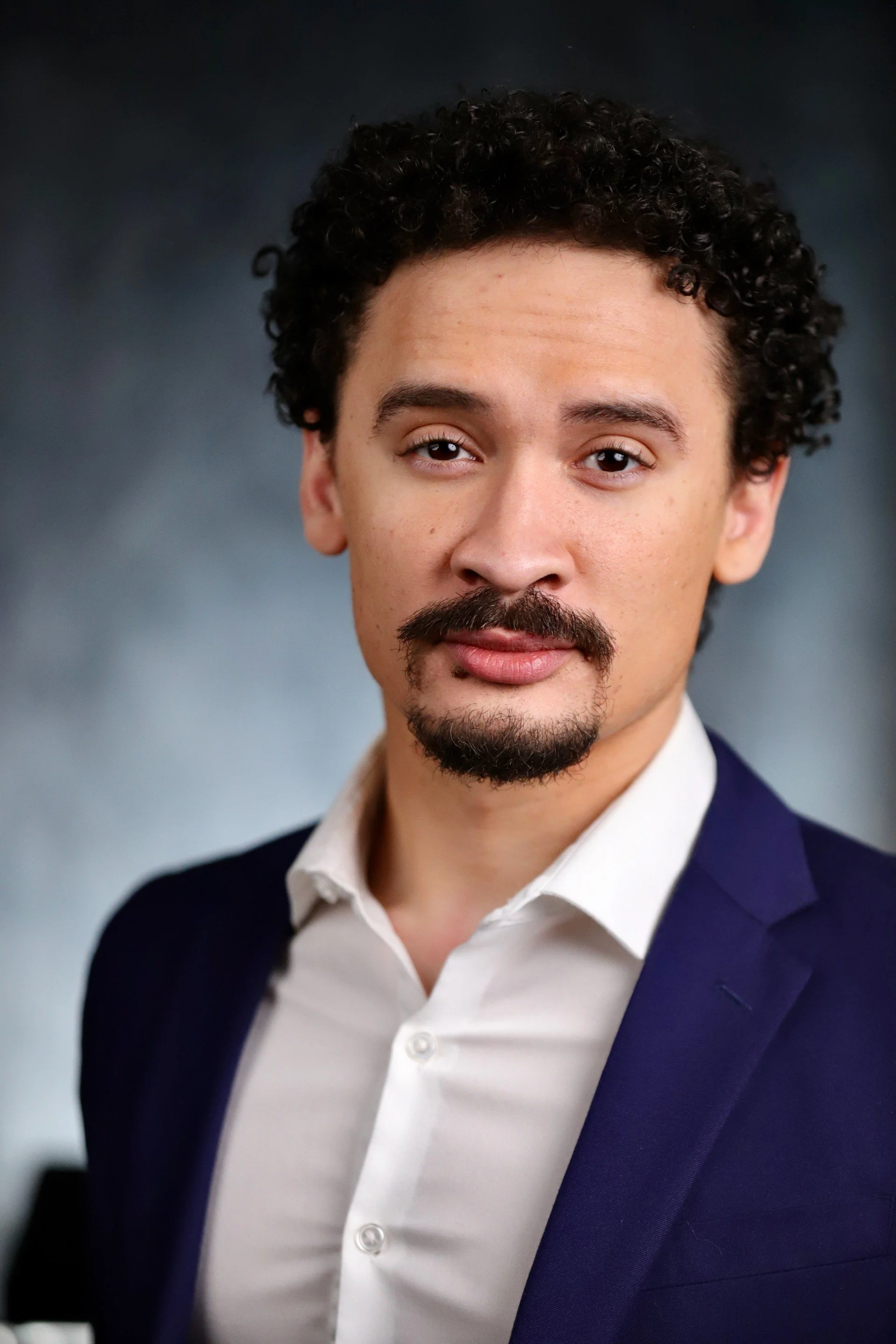 Headshot of a man with curly dark hair, a mustache, and goatee, dressed in a white shirt and navy blazer, against a blurred dark background.