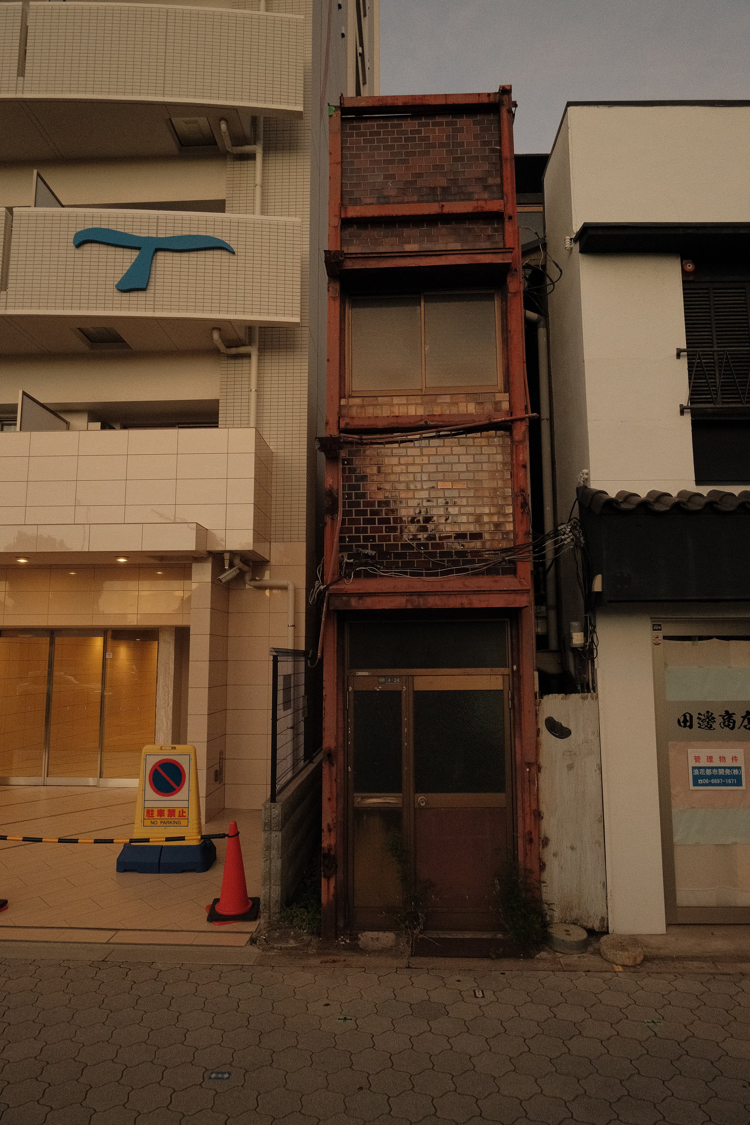 Old, unused elevator shaft with brick and metal framing in an urban setting, next to a modern building. The area is dimly lit, and there are traffic cones and a no parking sign in the foreground.