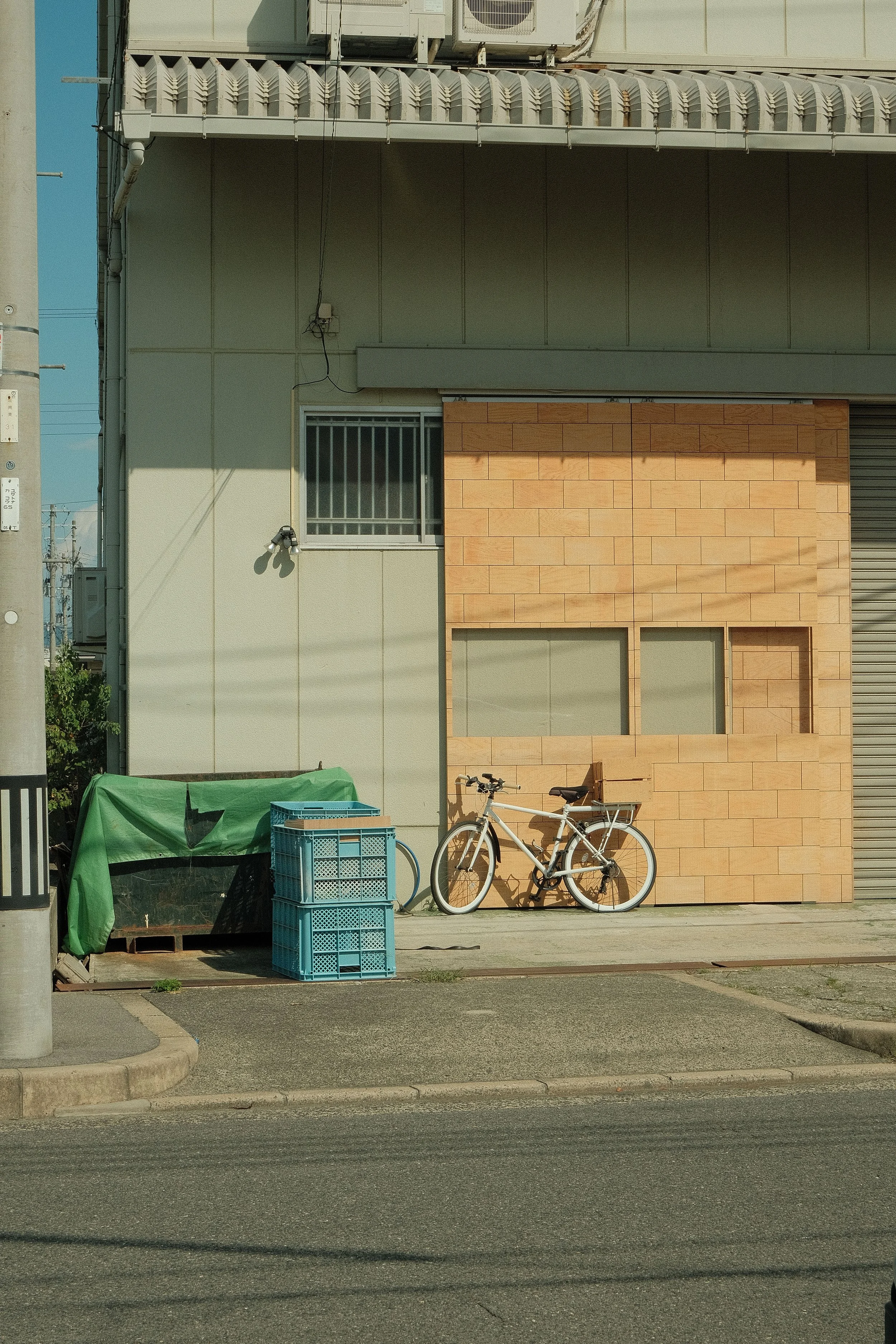 A beige building with a barred window, a security camera, and a partially visible garage door. In front, there is a bicycle, some blue crates, and a green tarp-covered object near a sidewalk and street.