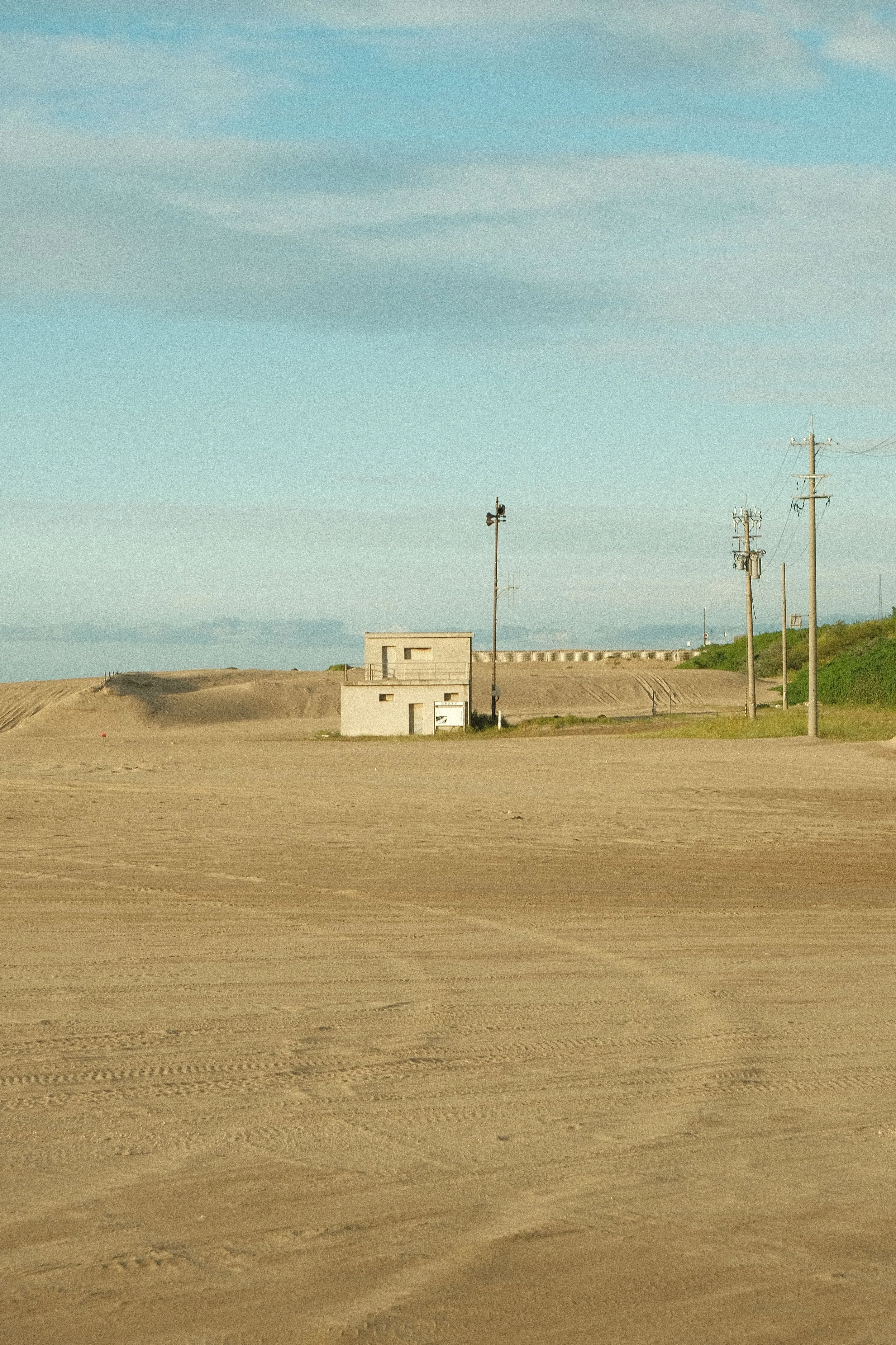 A small, white building with a flat roof and a wire fence in a barren, sandy landscape under a partly cloudy sky. Power poles and lines run along the right side of the image.