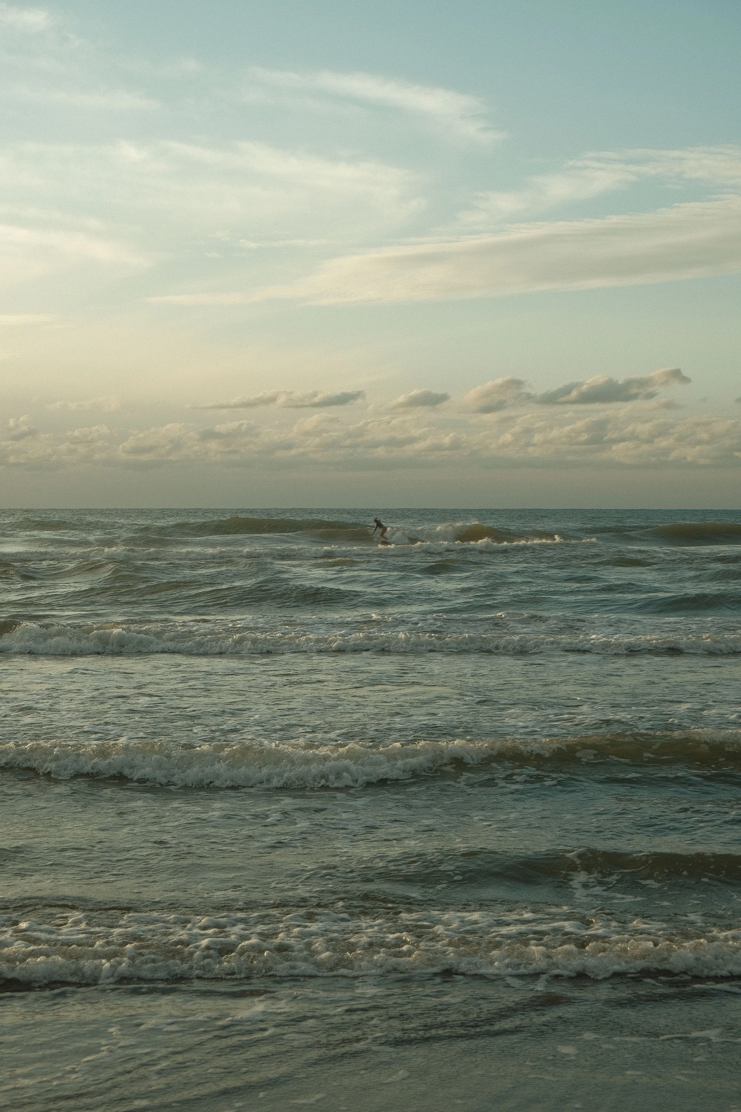 A calm ocean scene with gentle waves and a surfer riding a wave, under a cloudy sky with soft light.