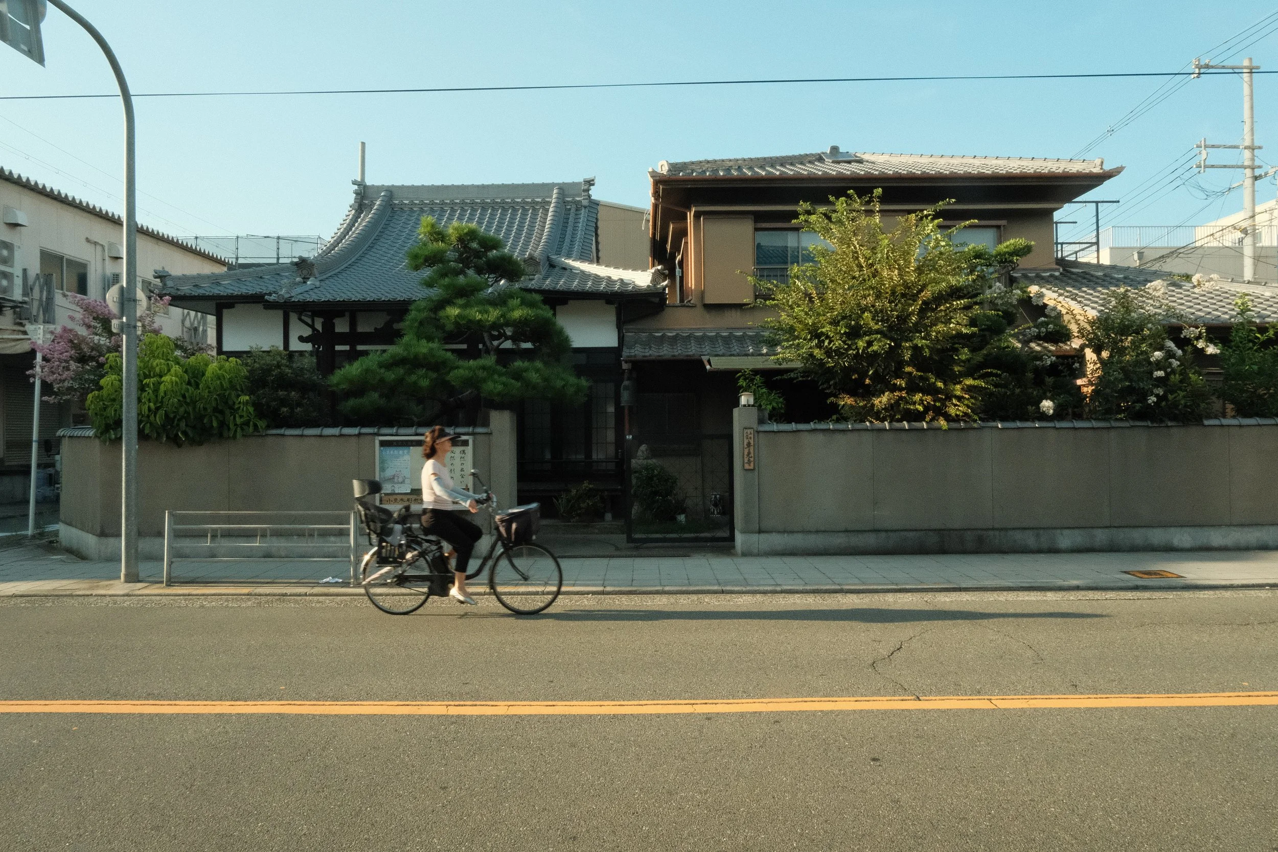 A woman riding a bicycle on the street in front of a traditional Japanese house with tiled roof and trees.