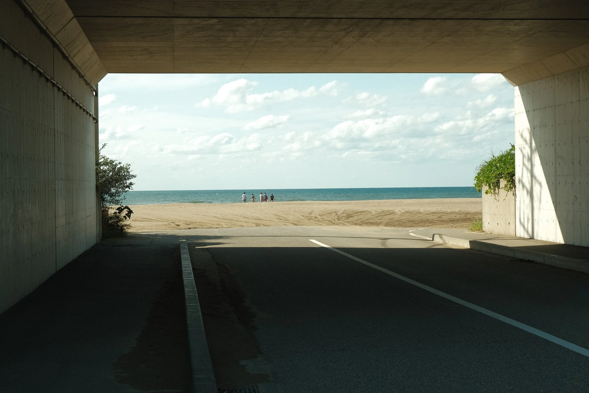View from under a bridge showing a sandy beach, the ocean, and a few people walking along the shoreline.
