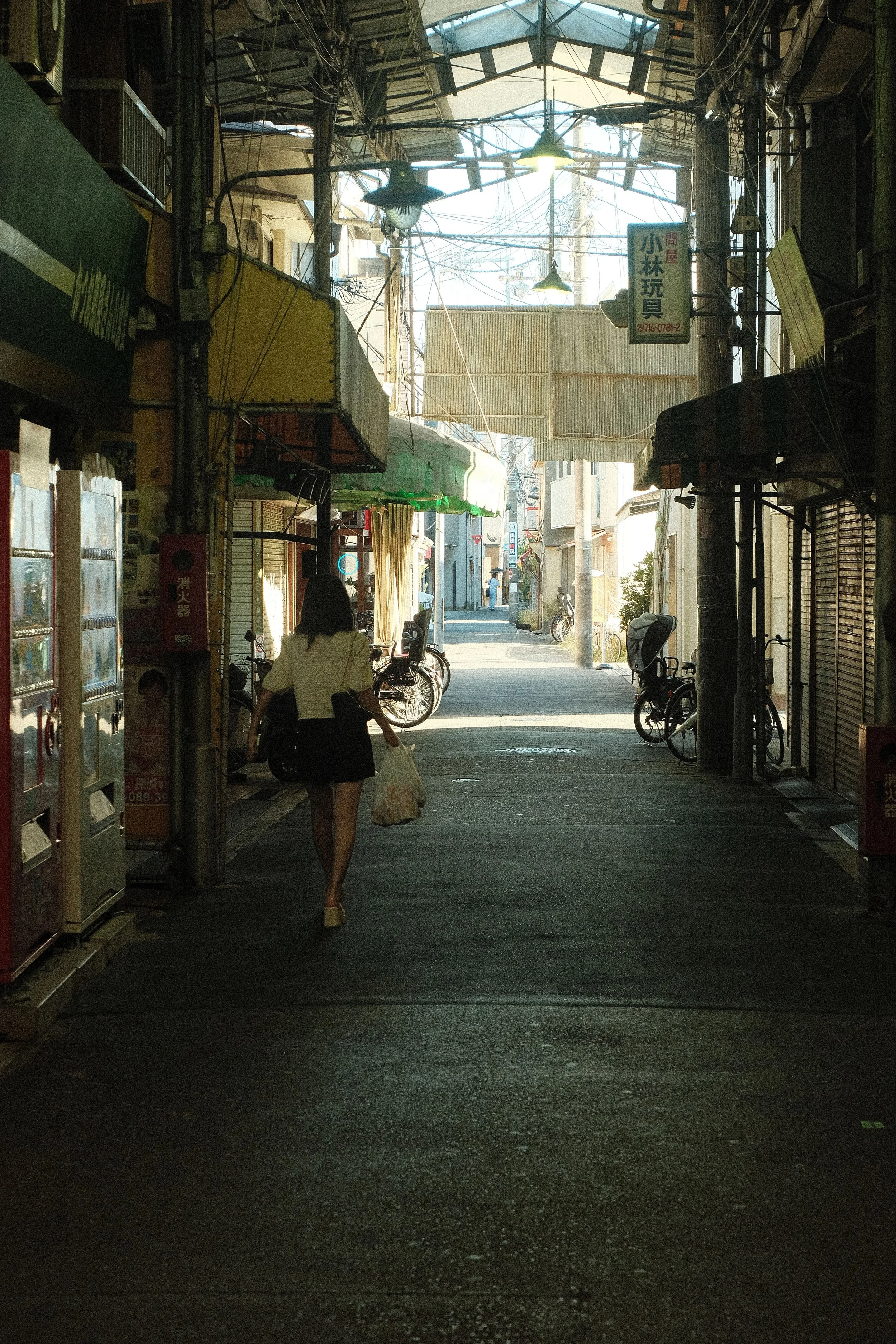 A woman walking through a covered outdoor market or shopping street, with bicycles and storefronts on either side, sunlight at the end of the street.
