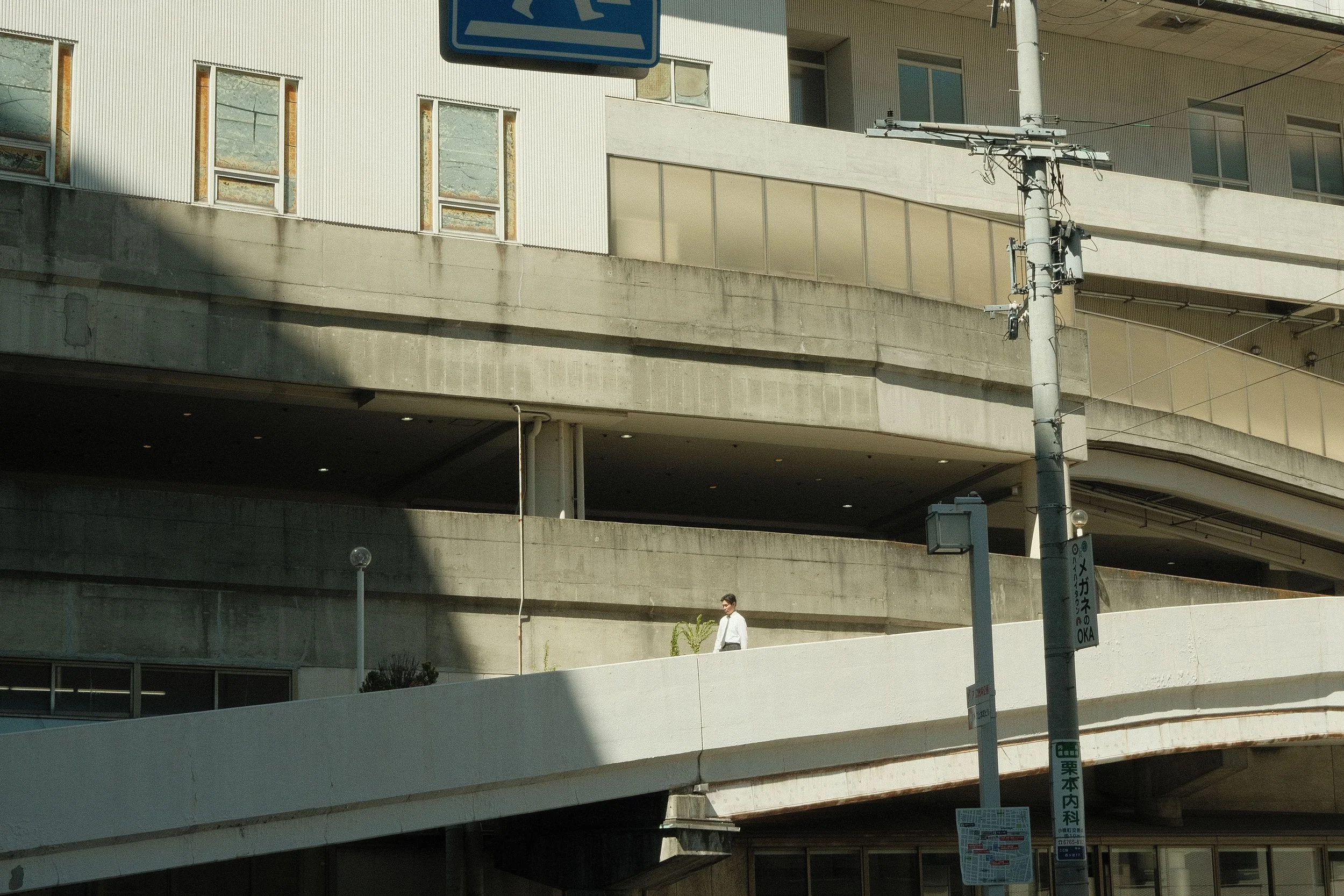 A city street scene featuring a multi-level parking garage with a concrete structure and multiple windows. There is a street sign hanging above and a utility pole with wires in front. A person is walking on the sidewalk below, and there are street la