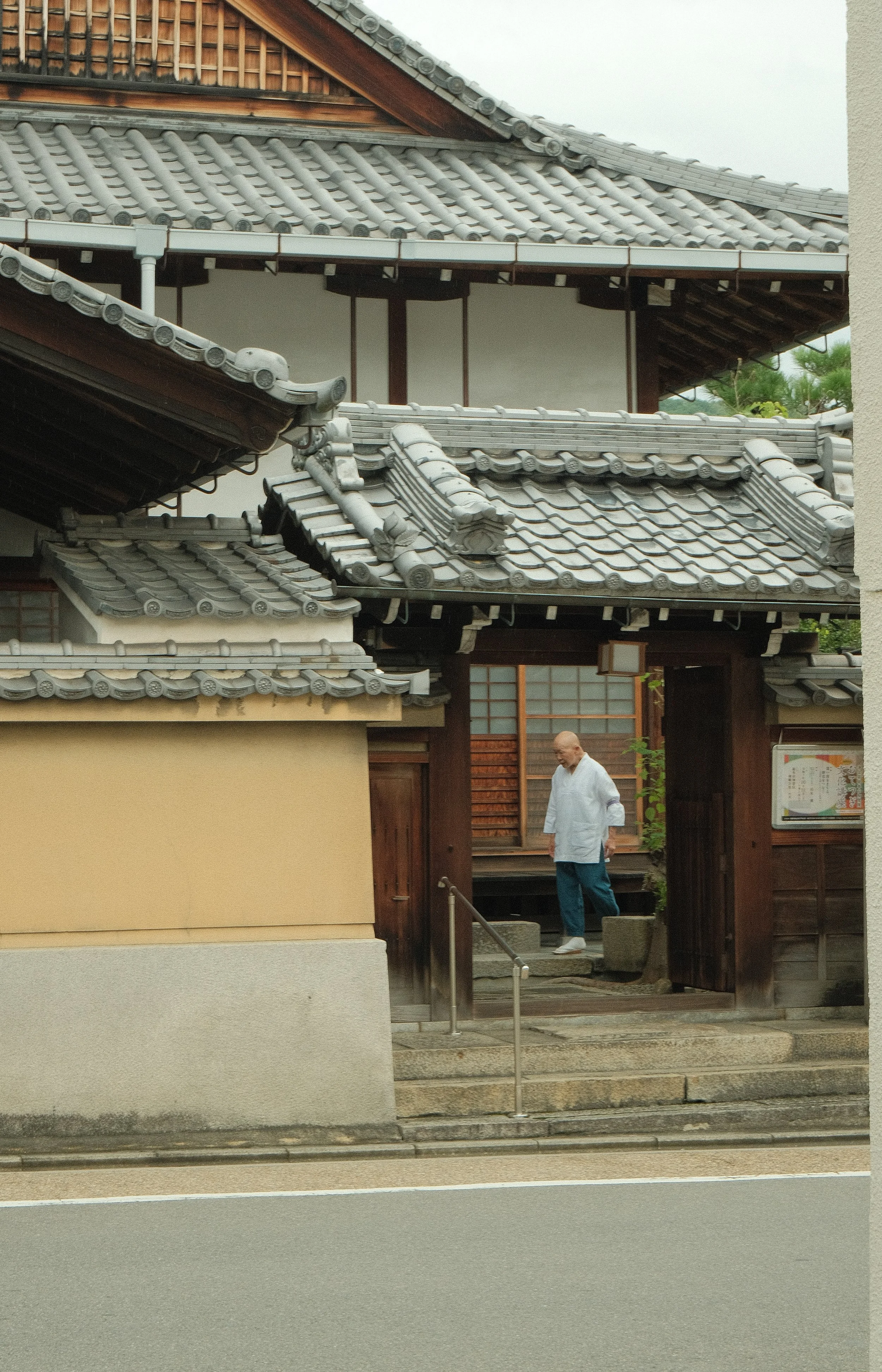 A man in traditional Japanese clothing stands at the entrance of a Japanese temple with tiled roofs and wooden structures.