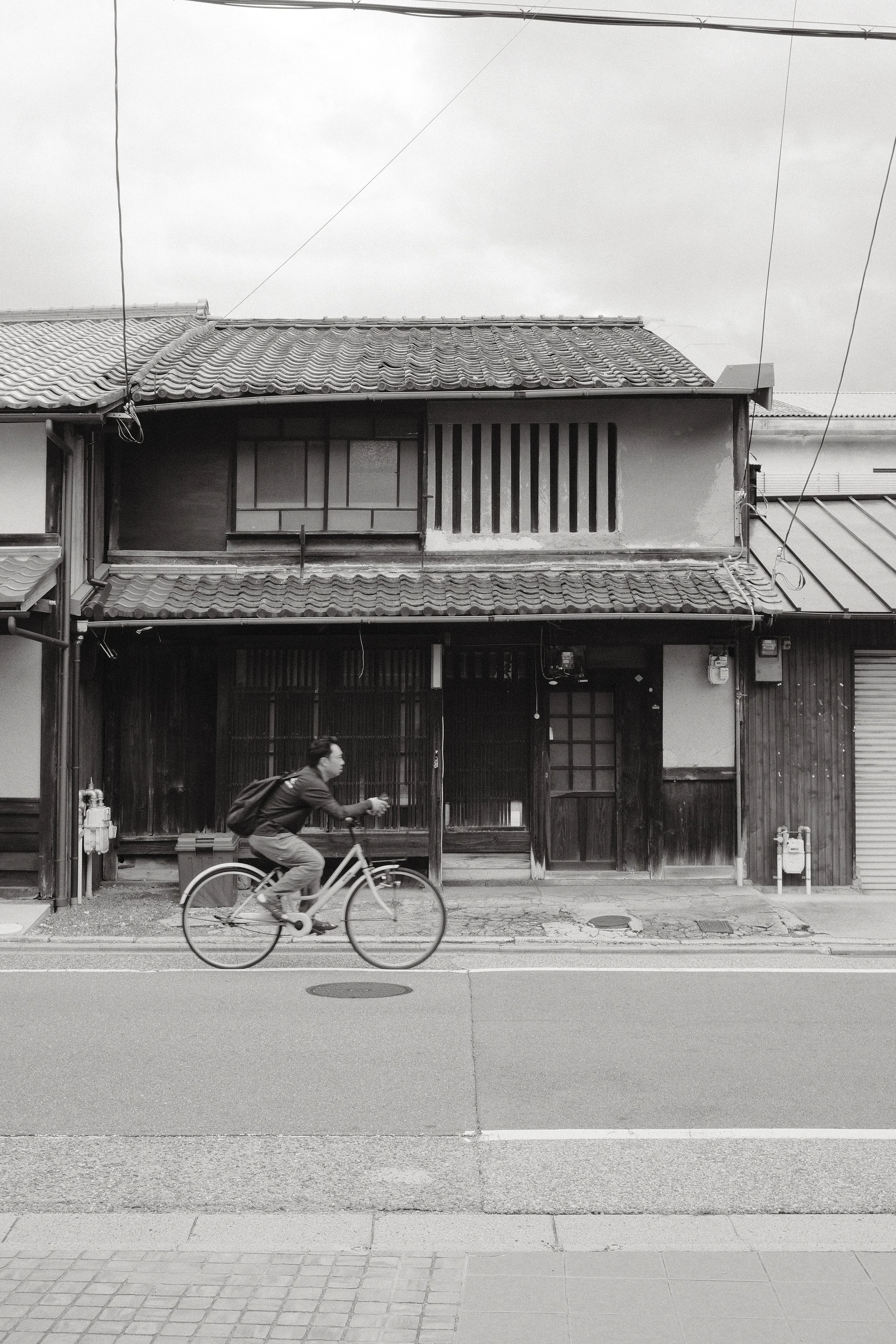 A man riding a bicycle on the street in front of a house with a tiled roof and a balcony with vertical bars.