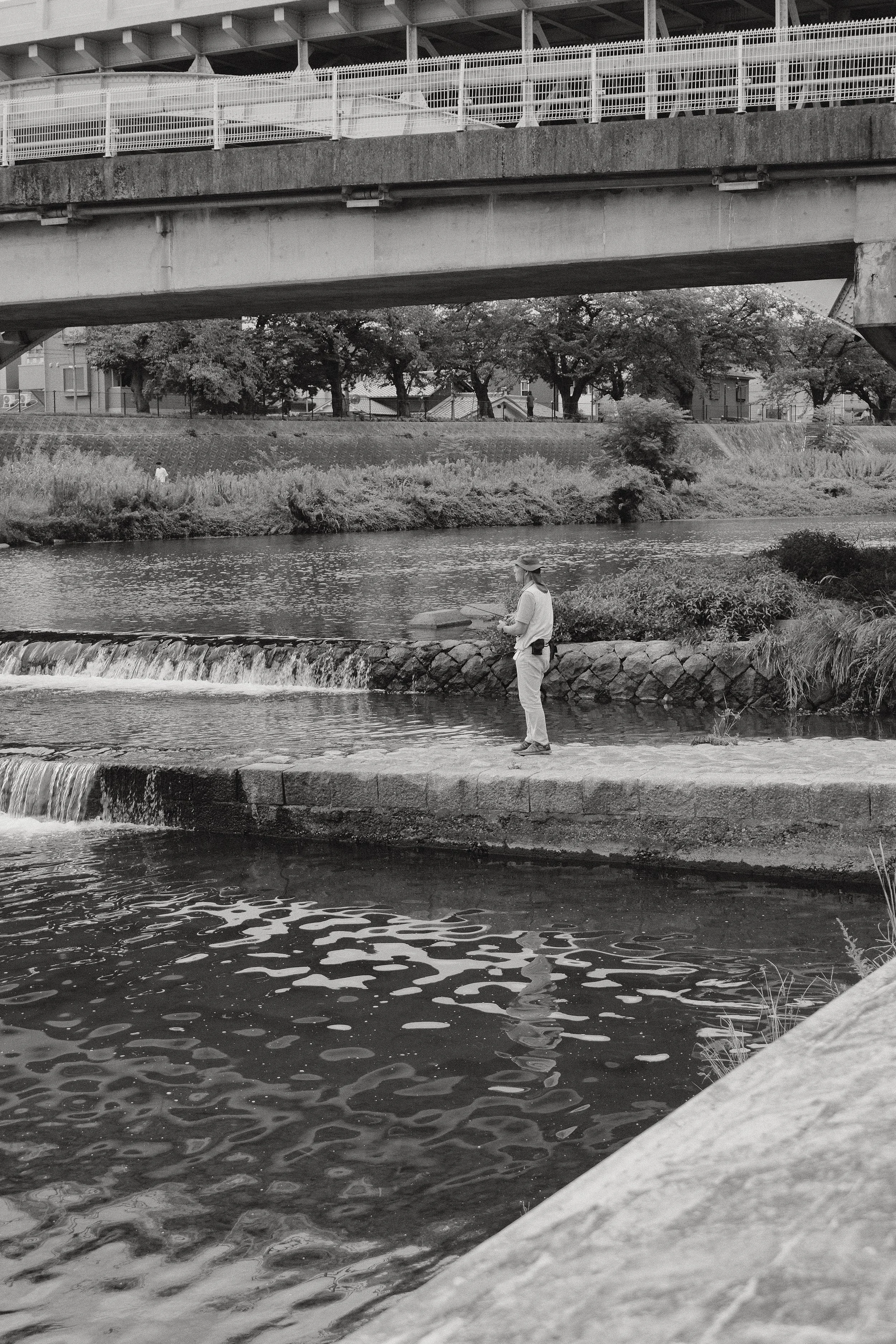 A person fishing by a river under a bridge with trees in the background, black and white photo.