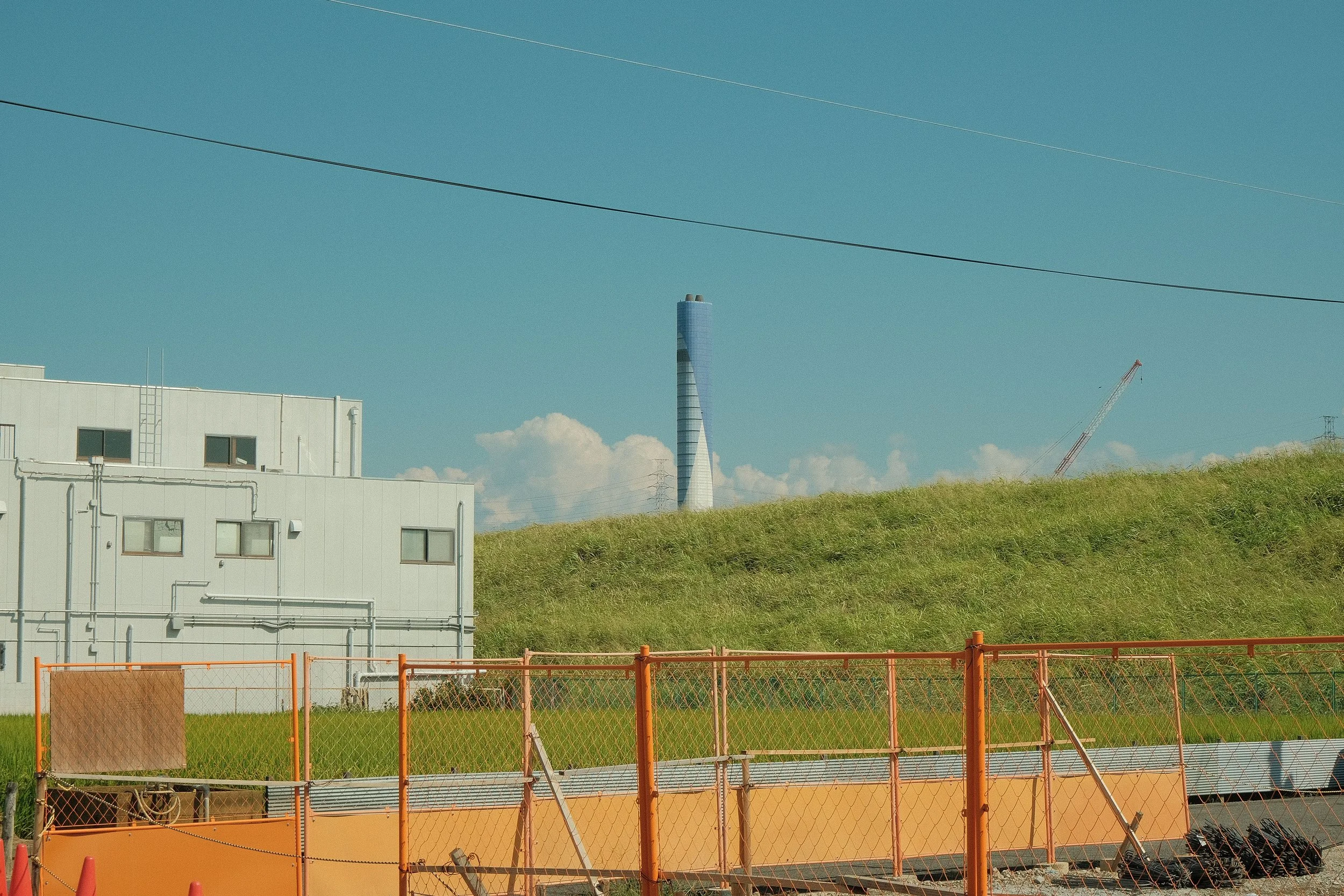 A construction site with a white building in the foreground, a grassy hill behind it, and a tall blue glass skyscraper in the background. There are power lines across the sky, and construction equipment including a crane on the right side.