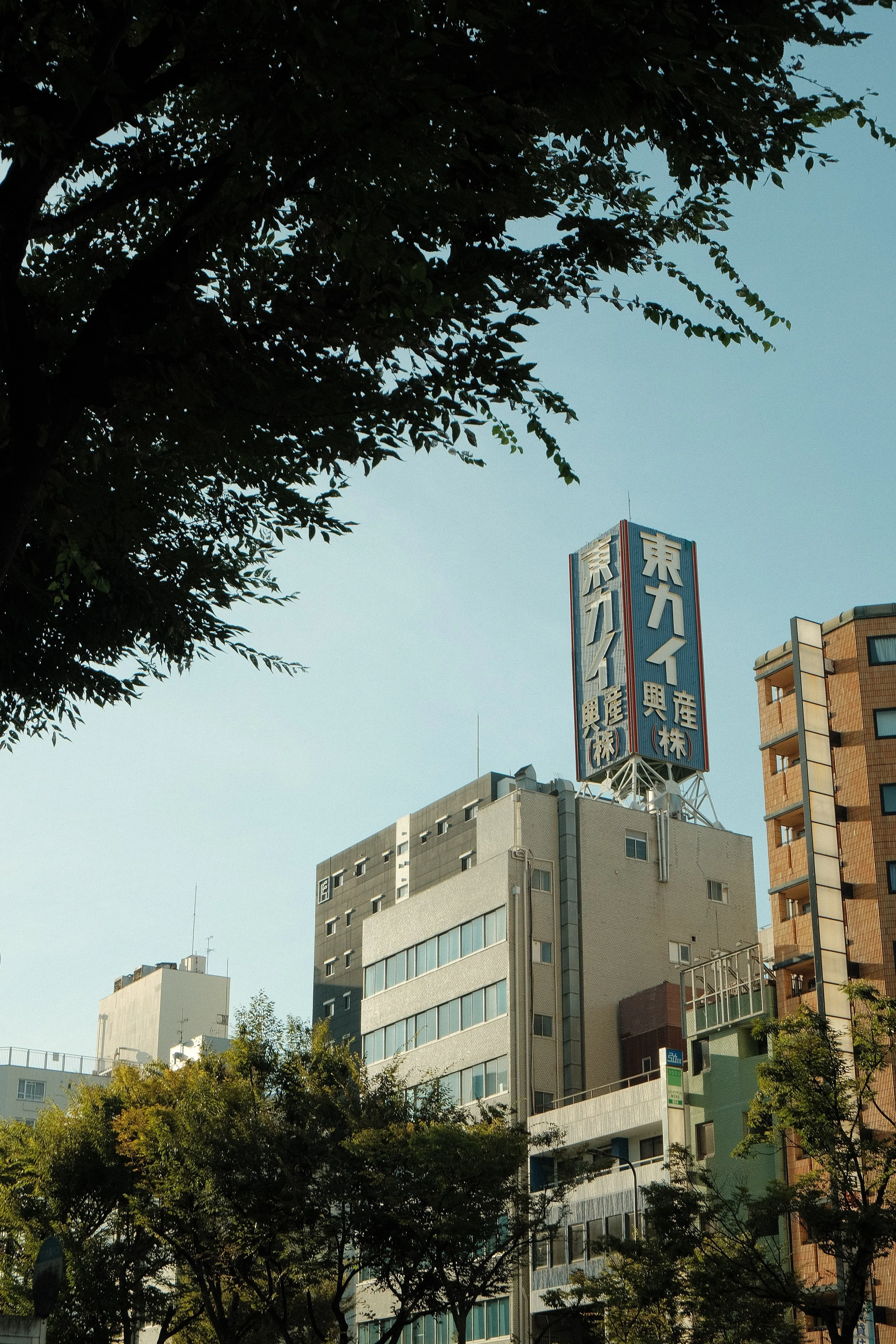 Cityscape with tall buildings and a blue sky, trees in the foreground, and a prominent sign with Japanese characters on the rooftop of one building.
