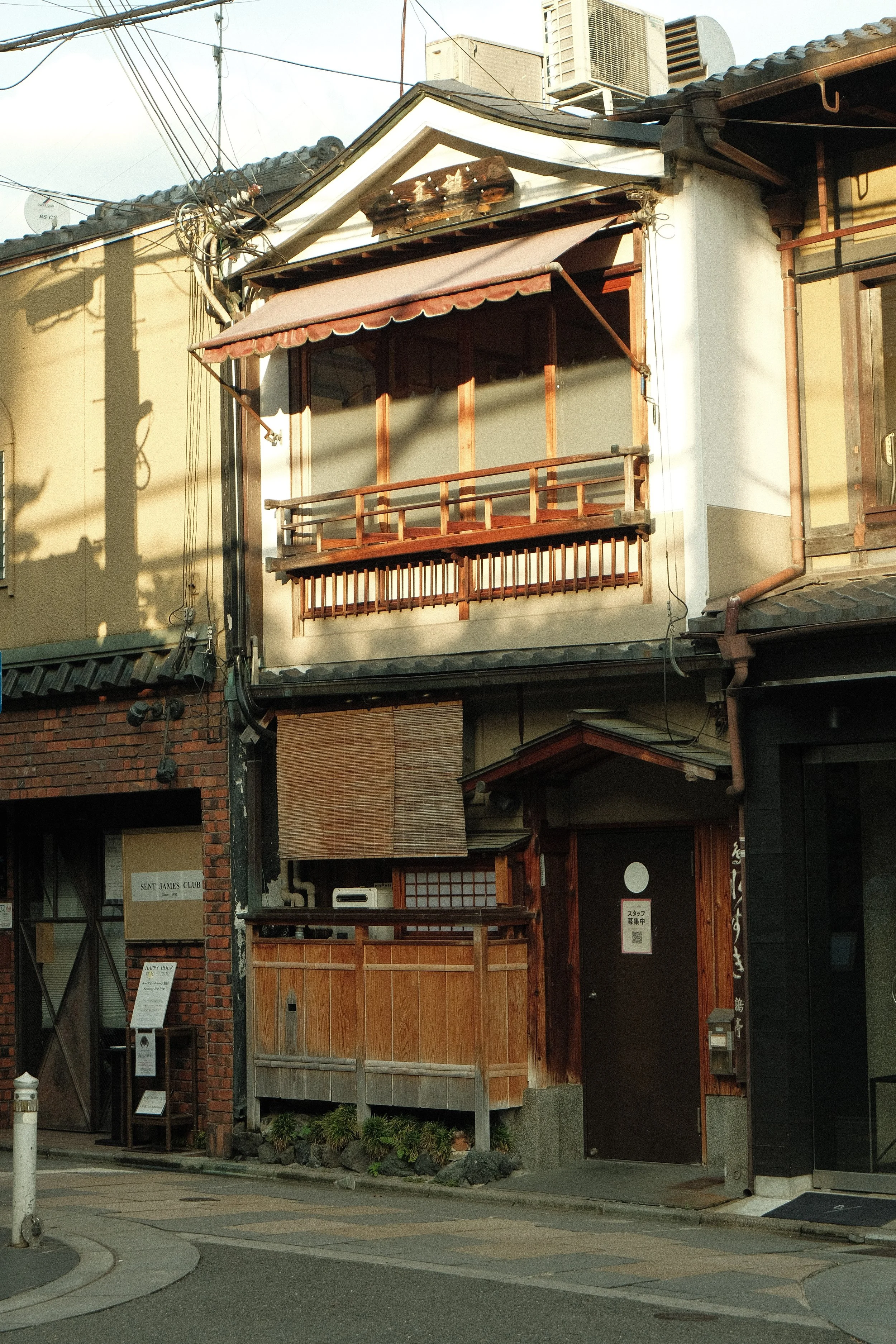 A traditional Japanese building with a wooden balcony and sliding doors, adjacent to other buildings, showing a modern cityscape in the background with utility poles and air conditioning units on the roof.