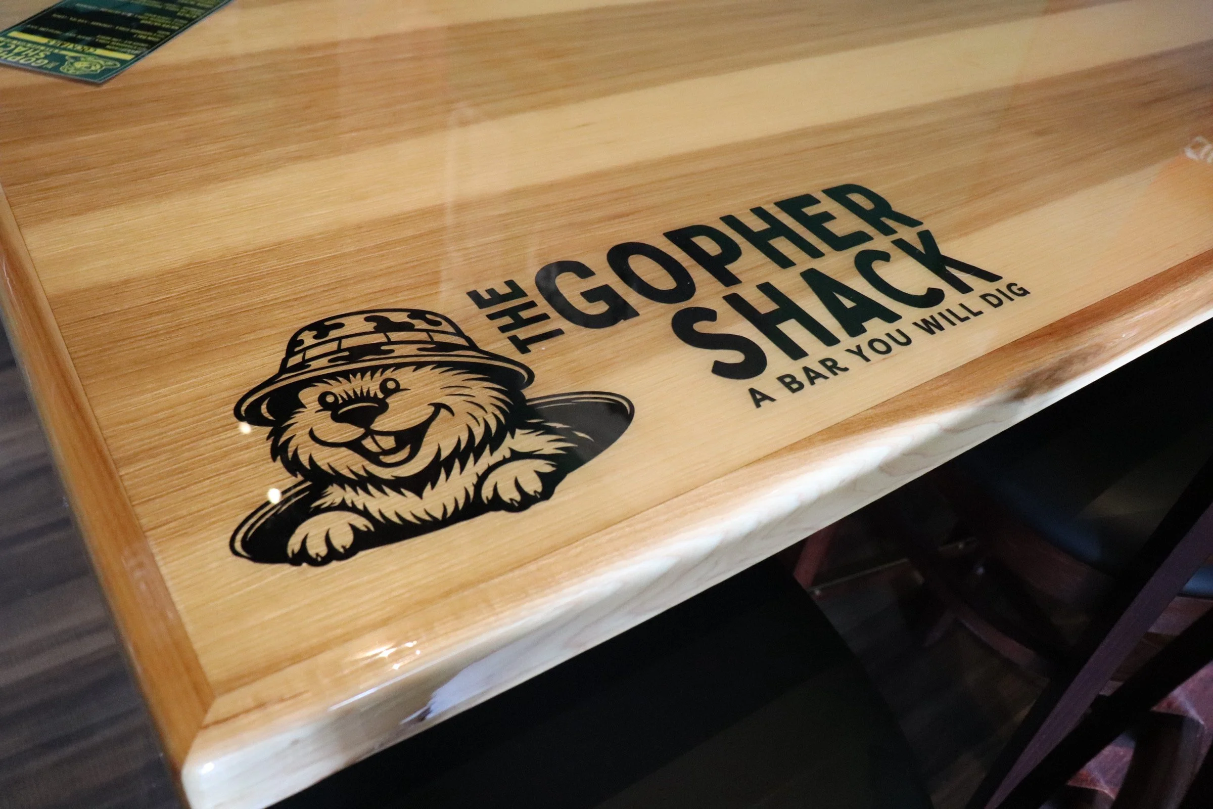 Wooden bar top with the logo and text 'The Gopher Shack' and the subtitle 'A Bar You Will Dig' along with a cartoon gopher wearing a hat.