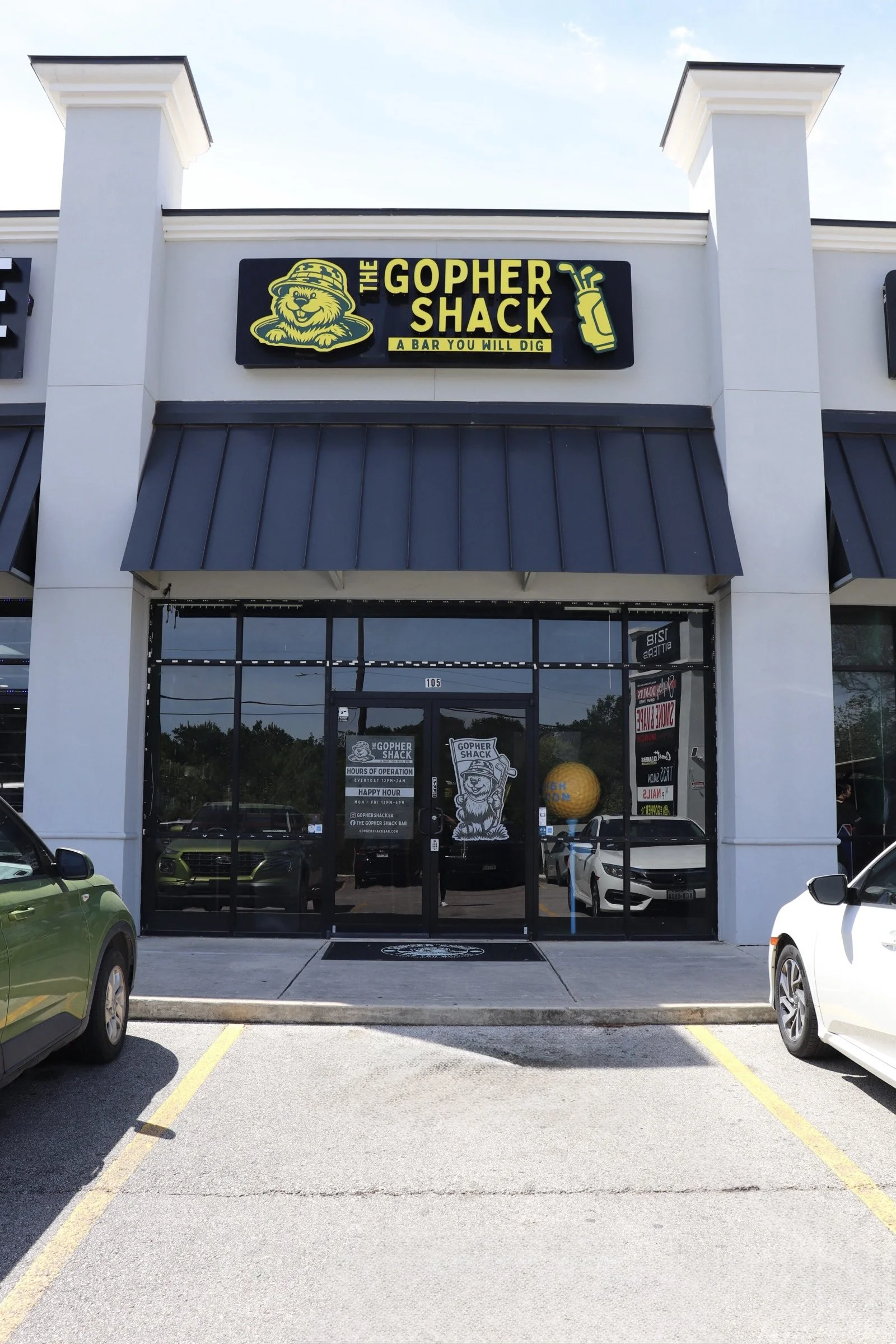 Front view of a Gopher Shack restaurant with a parking lot in foreground. The sign features a gopher cartoon wearing a hat, and text that reads 'The Gopher Shack' with a tagline 'A Bar You Will Dig.' The entrance has glass doors and windows, and cars are parked on either side.