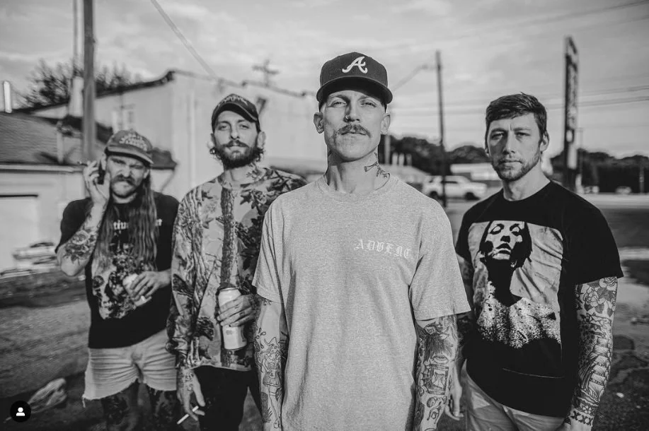 Four men standing outdoors in a line, with two in the front and two behind, in an urban setting with power lines and buildings in the background. They have tattoos, and two are wearing caps. The photo appears to be taken during the daytime.