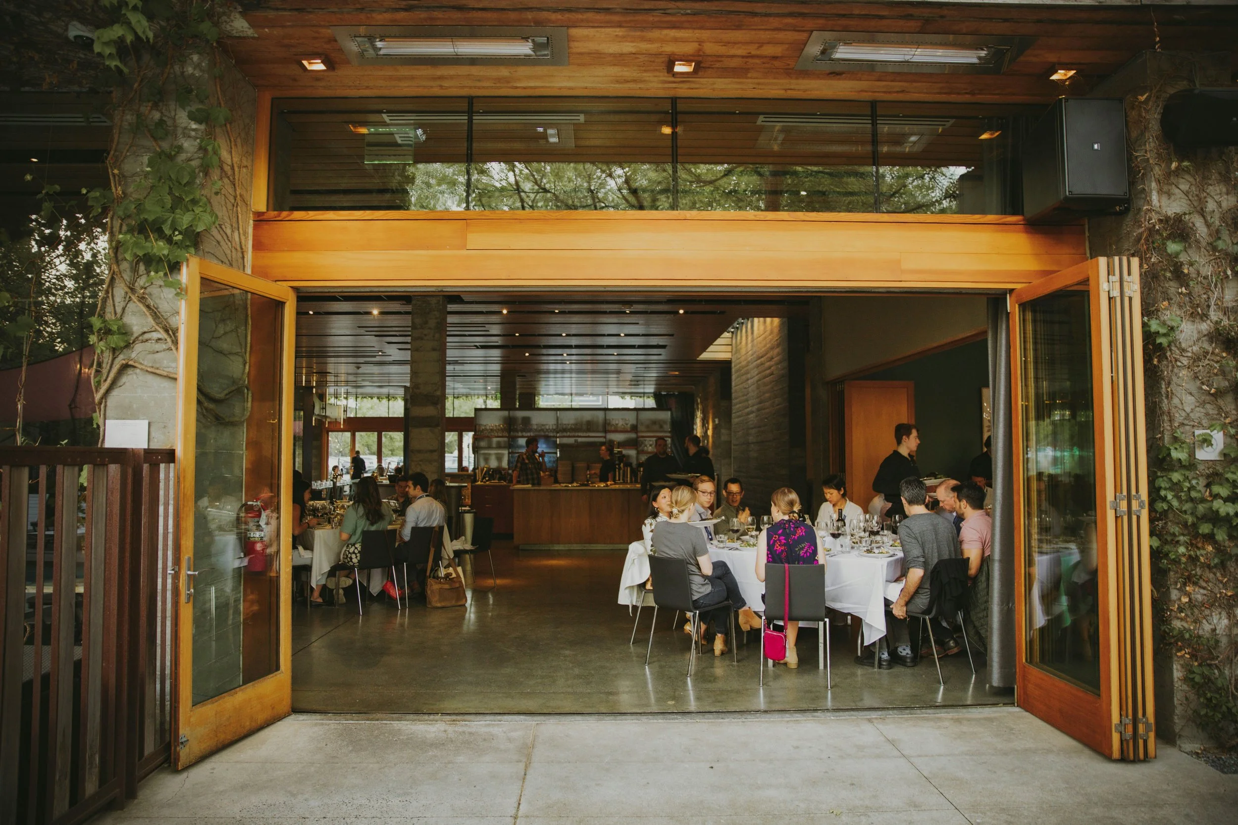 People dining inside a restaurant, viewed through open wooden-framed glass doors, with some seated at tables and others ordering at the counter, in a modern space with wooden accents and greenery outside.