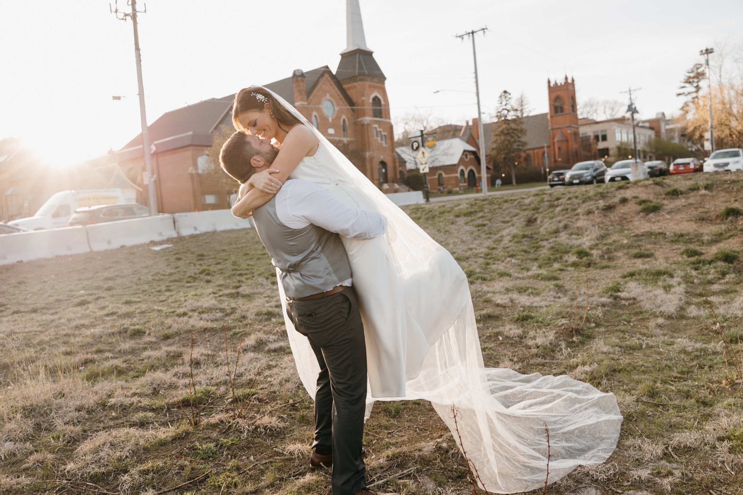 A newlywed couple; the groom lifting the bride in a field during sunset, with church and houses in background.