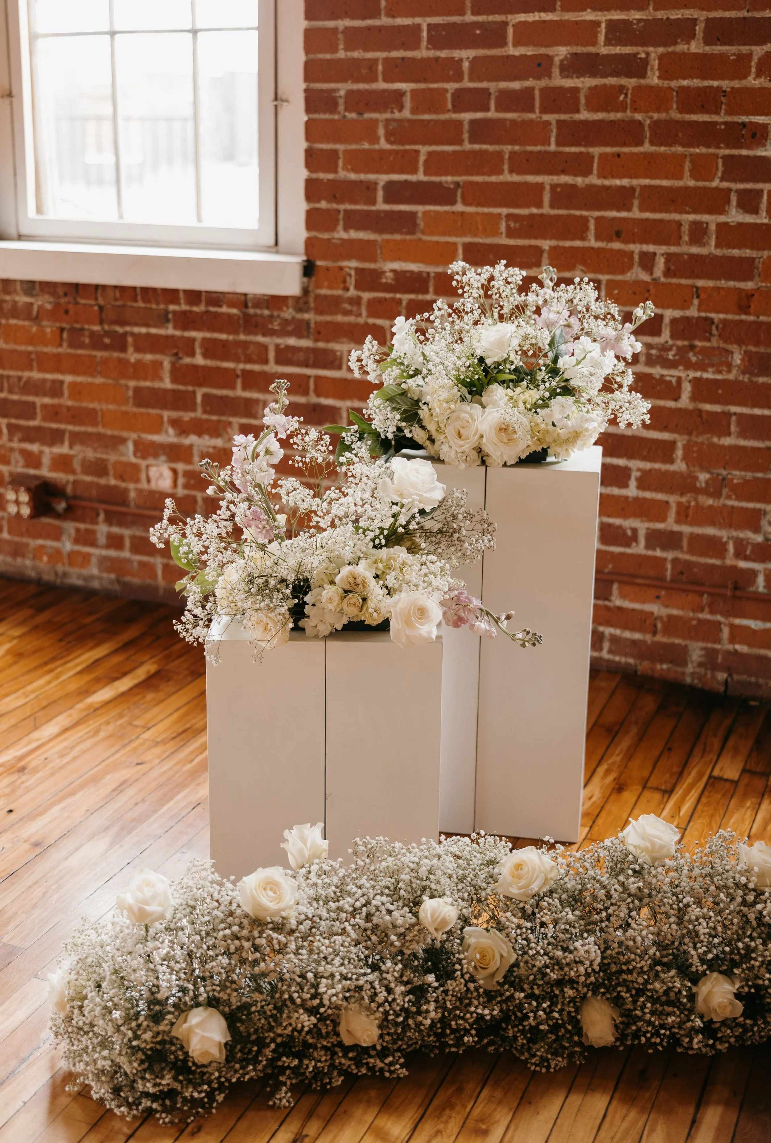 White floral arrangements with roses and baby's breath on two white platforms and on the floor in front of a brick wall and wooden floor near a window.