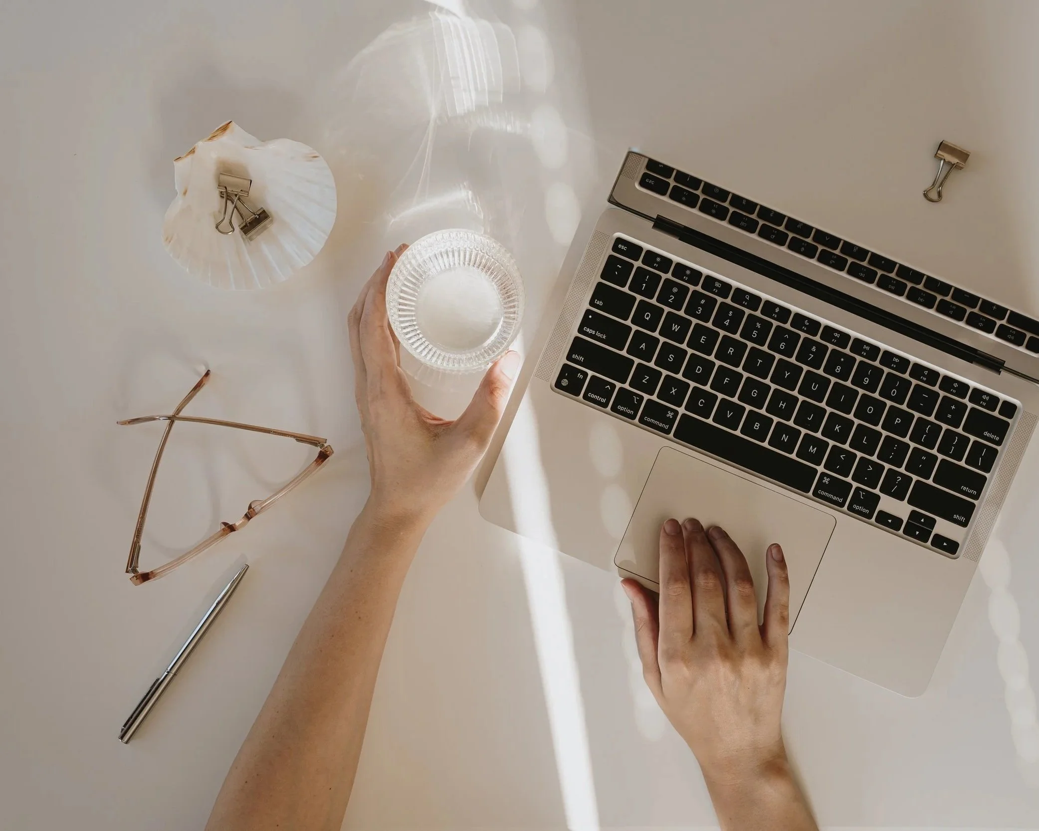 Overhead view of a workspace with a person's hands typing on a silver laptop, holding a small glass container, and surrounded by a pair of glasses, a pen, a paperclip, and a light-colored paper cupcake wrapper on a white table.