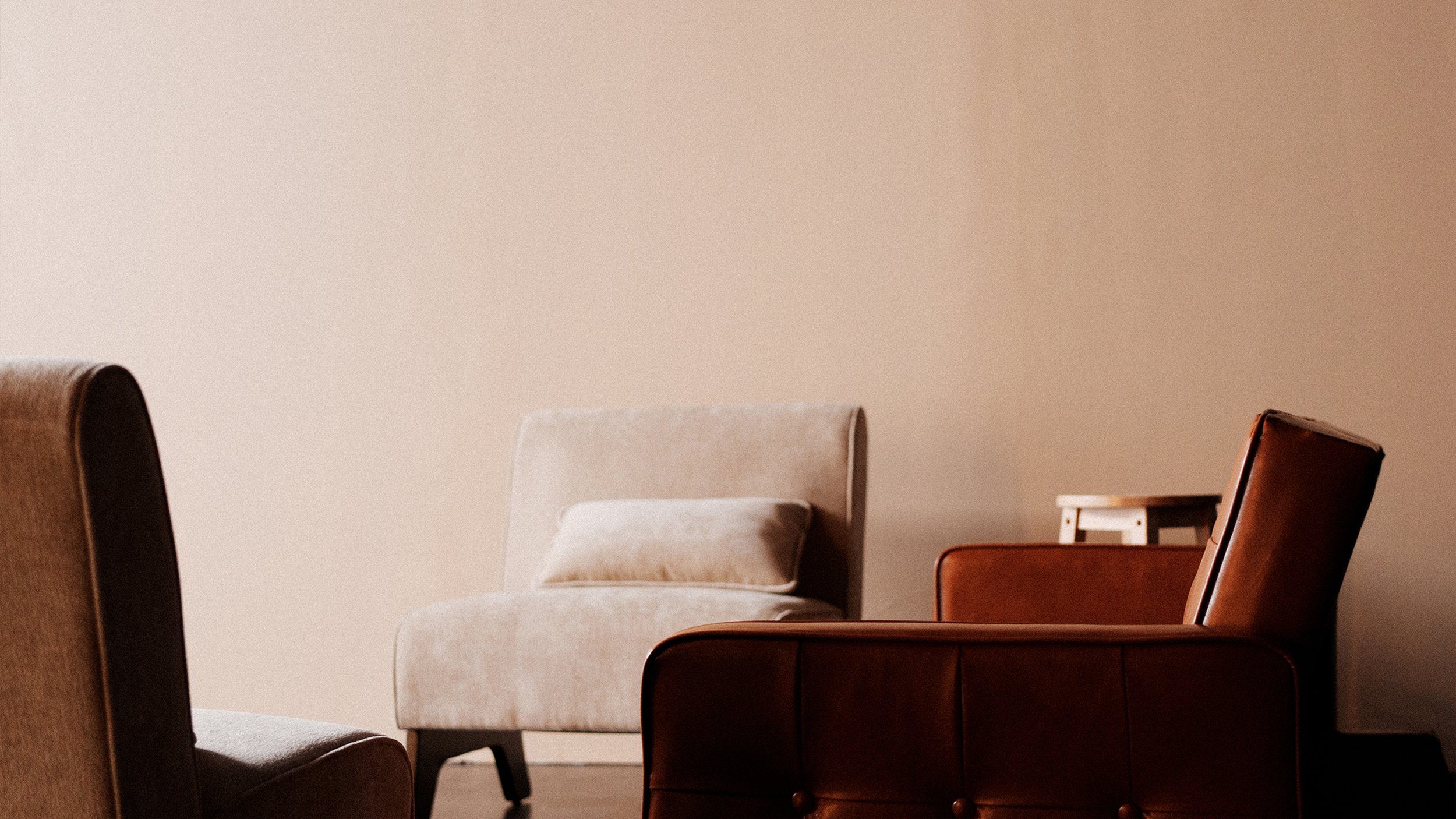 A minimalist living room with beige, brown, and tan chairs and a small side table, set against a plain beige wall.